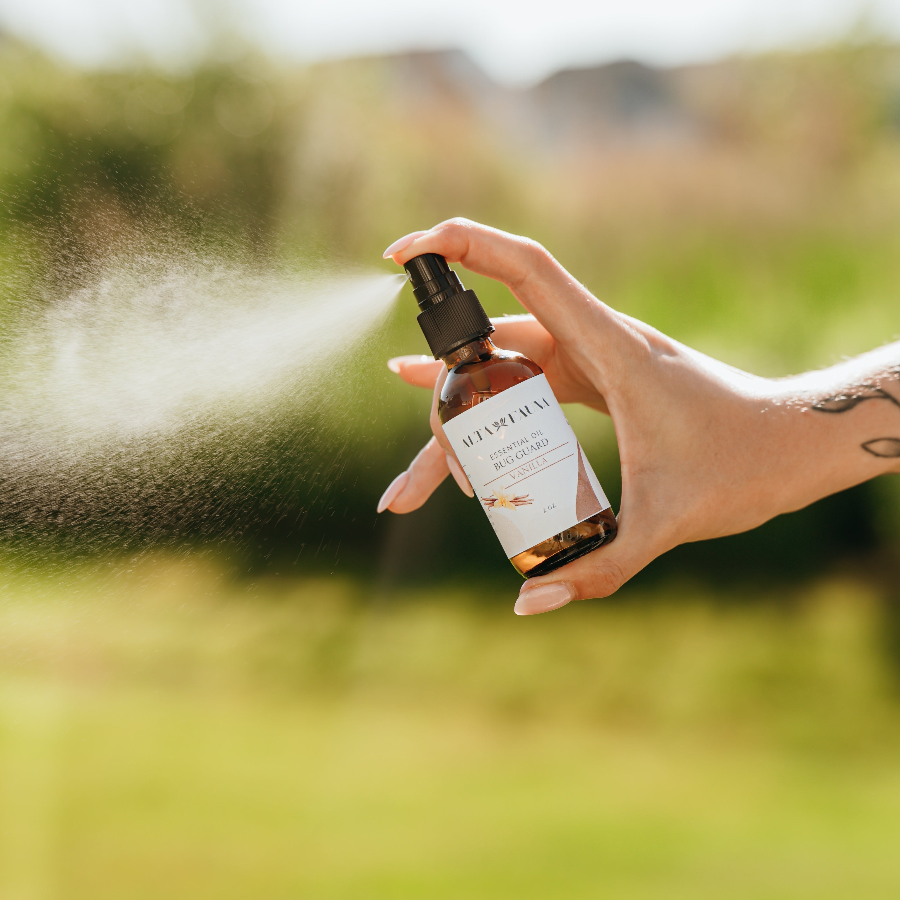Alta Fauna Vanilla Bug Guard being held by a hand in an outdoor setting with a fine mist coming from the amber, glass spray bottle.