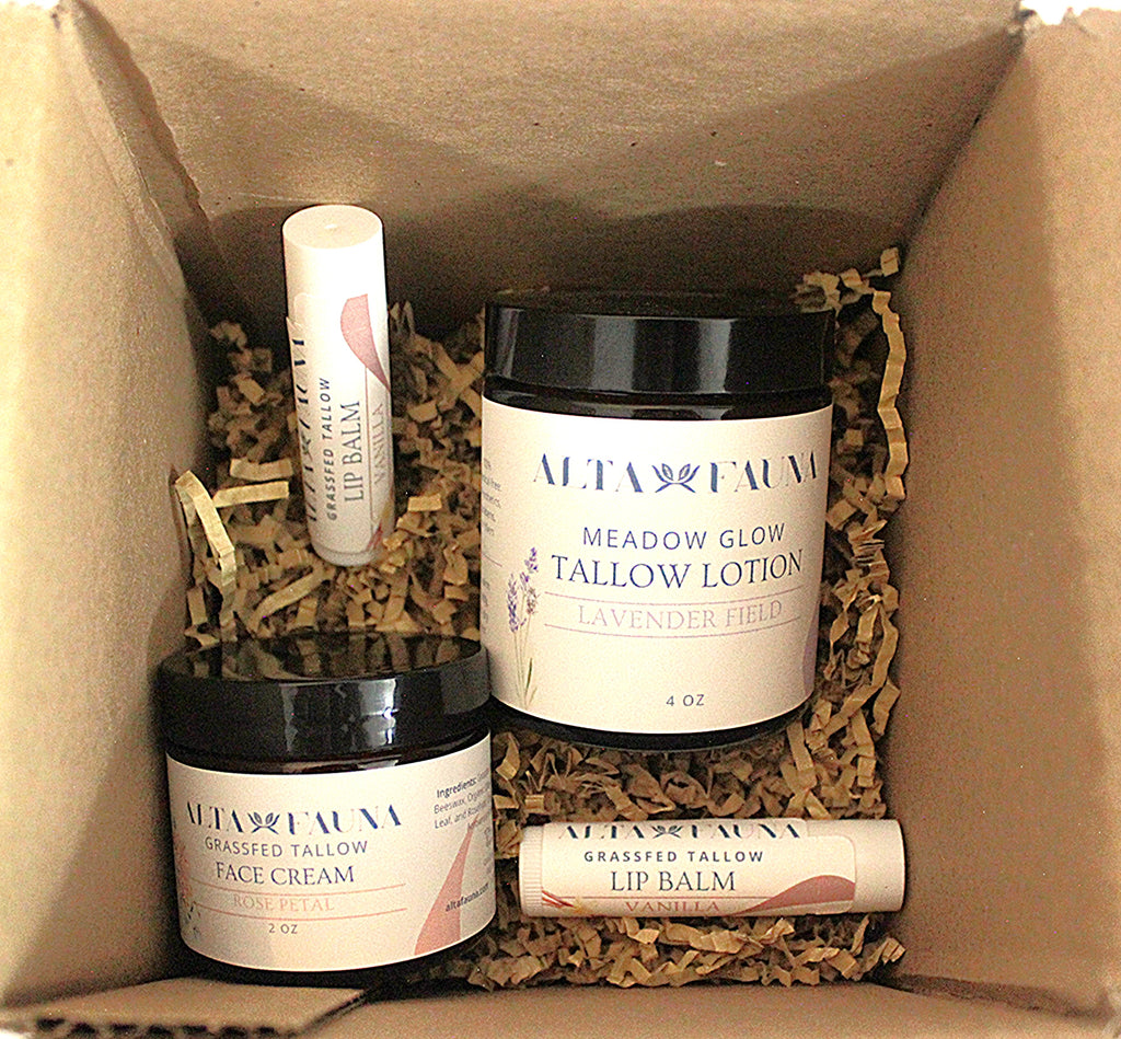 A boxed set of Alta Fauna natural skincare products nestled in brown paper shred inside a cardboard box. The set includes Meadow Glow Tallow Lotion in lavender field, Grassed Tallow Face Cream, and two lip balms.