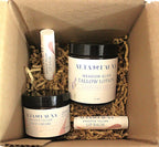 A boxed set of Alta Fauna natural skincare products nestled in brown paper shred inside a cardboard box. The set includes Meadow Glow Tallow Lotion in lavender field, Grassed Tallow Face Cream, and two lip balms.