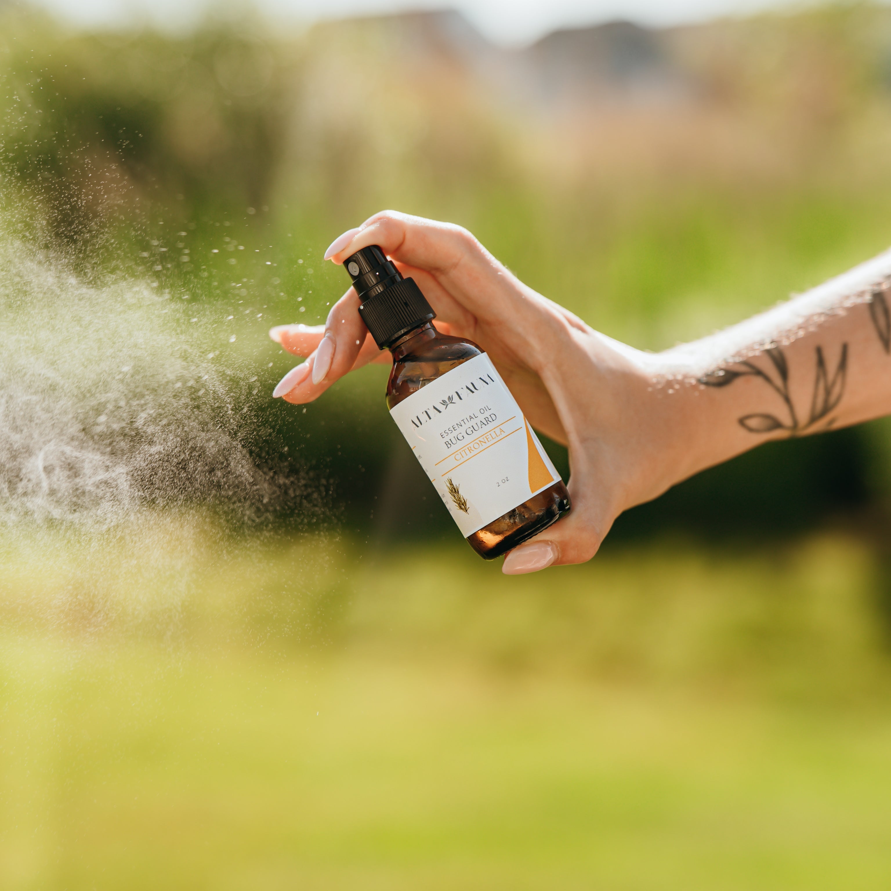 Alta Fauna Citronella Bug Guard being held by a hand in an outdoor setting with a fine mist coming from the amber, glass spray bottle.