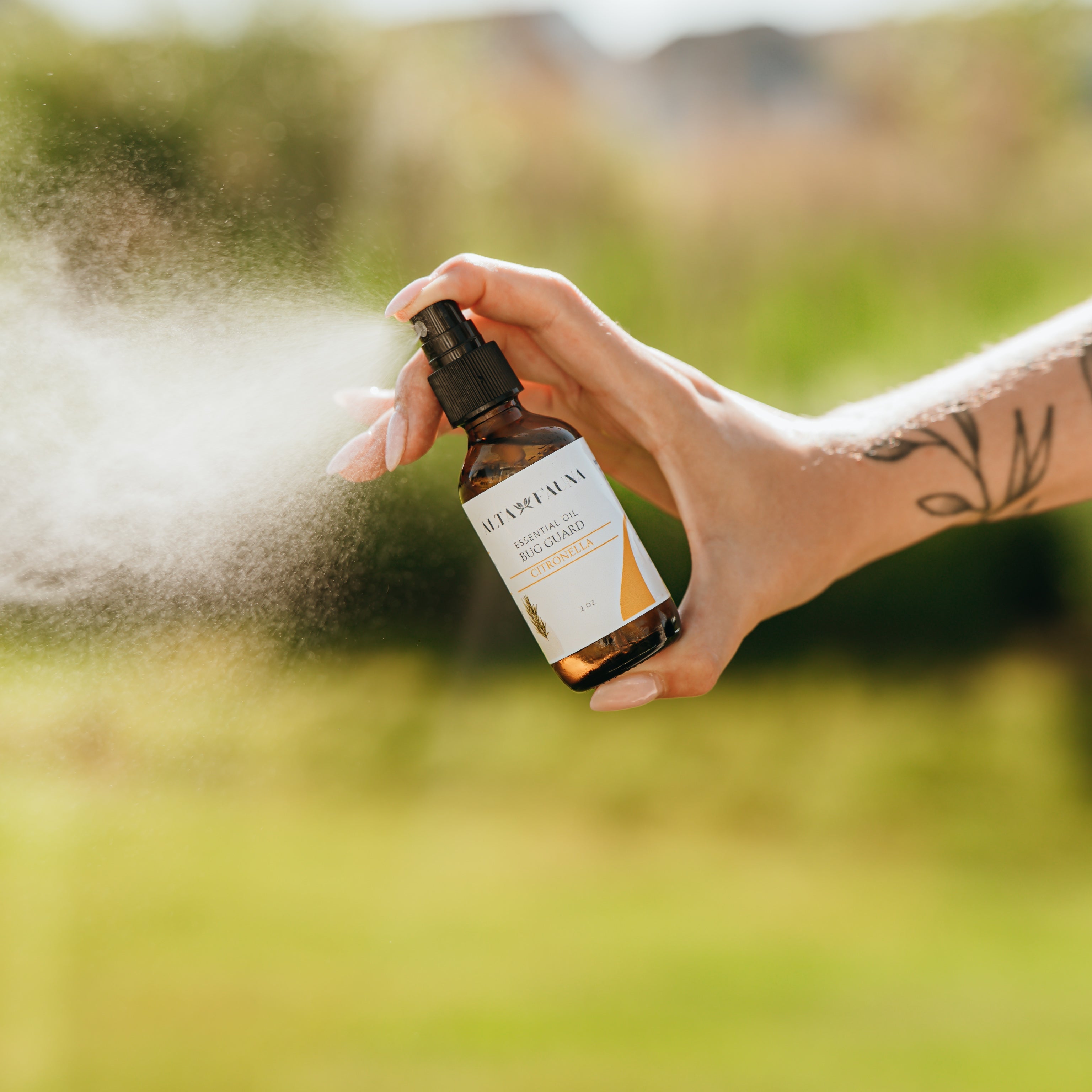 Alta Fauna Citronella Bug Guard being held by a hand in an outdoor setting with a fine mist coming from the amber, glass spray bottle.