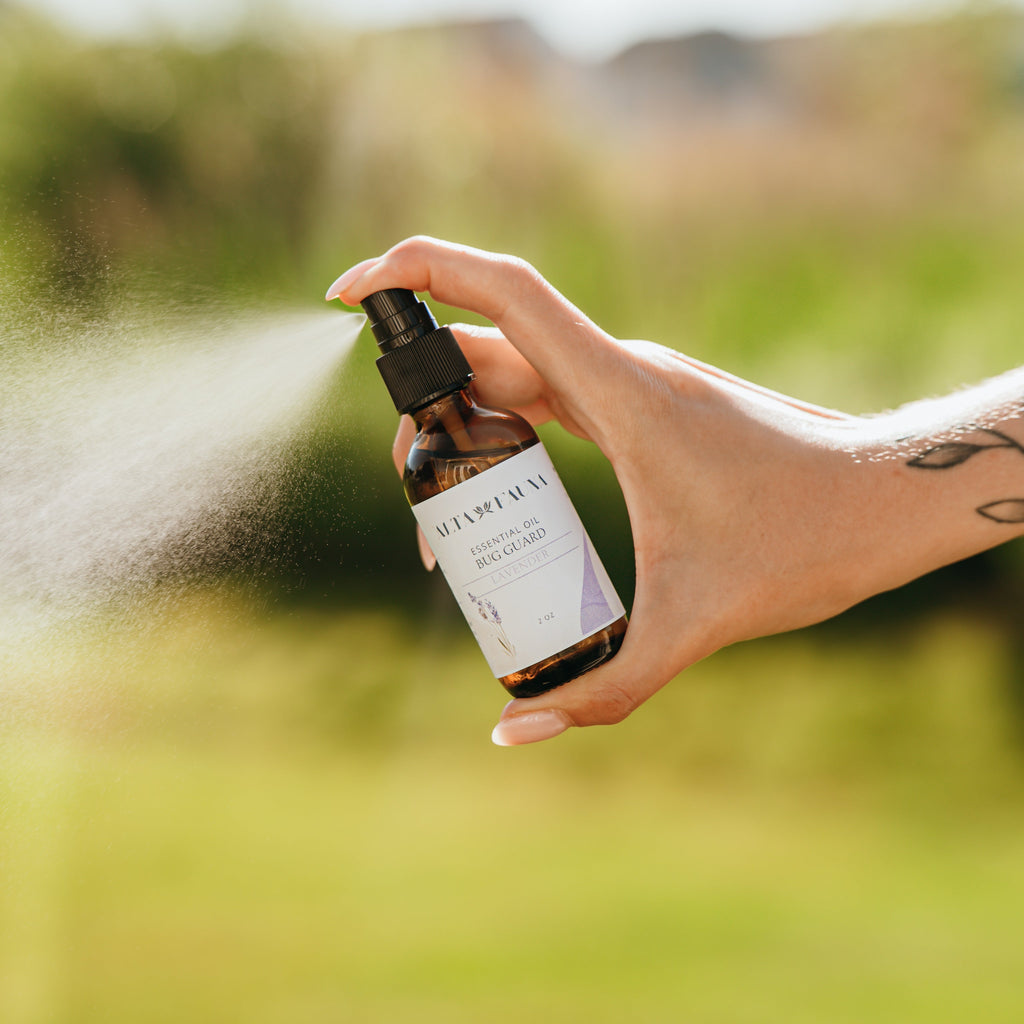 Alta Fauna Lavender Bug Guard being held by a hand in an outdoor setting with a fine mist coming from the amber, glass spray bottle.