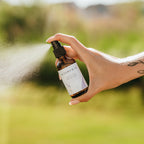 Alta Fauna Lavender Bug Guard being held by a hand in an outdoor setting with a fine mist coming from the amber, glass spray bottle.