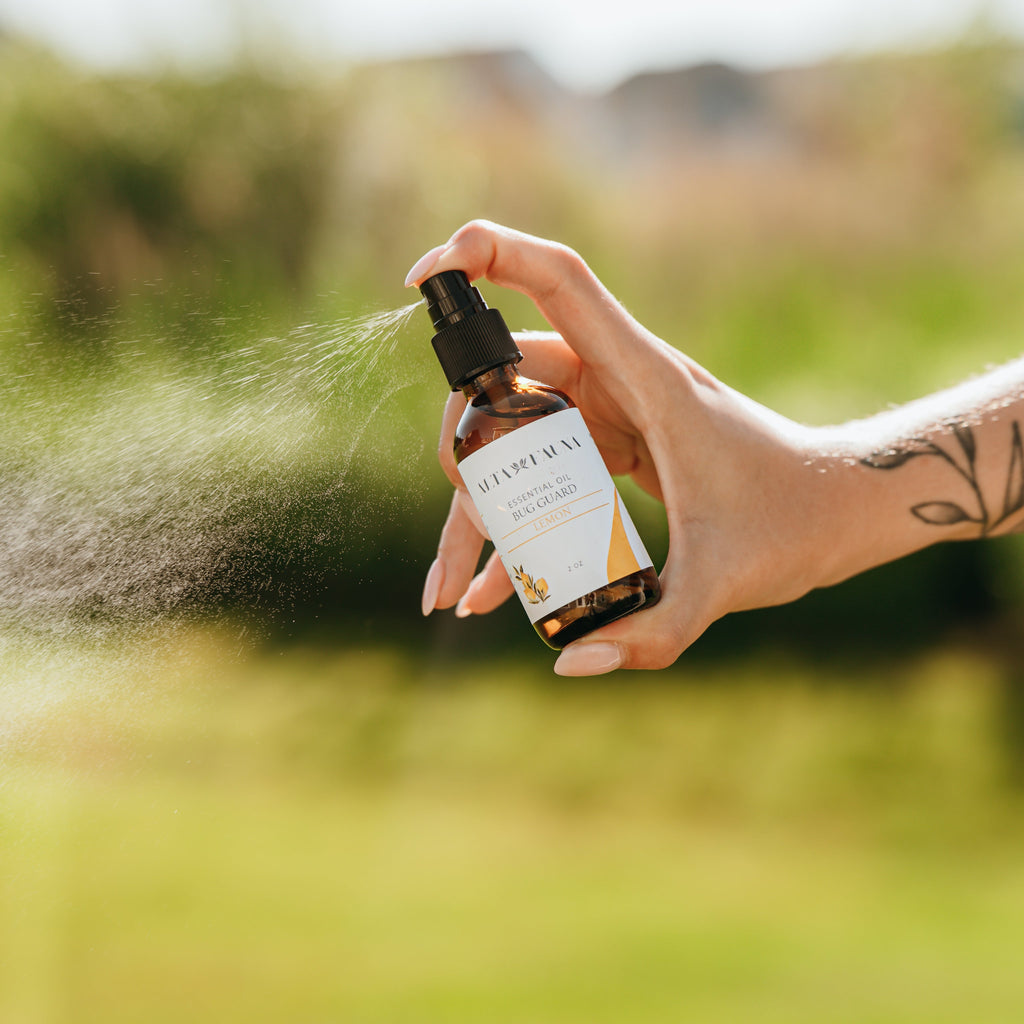 Alta Fauna Lemon Bug Guard being held by a hand in an outdoor setting. A fine mist is coming from the amber, glass spray bottle. 