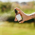 Alta Fauna Lemon Bug Guard being held by a hand in an outdoor setting. A fine mist is coming from the amber, glass spray bottle. 