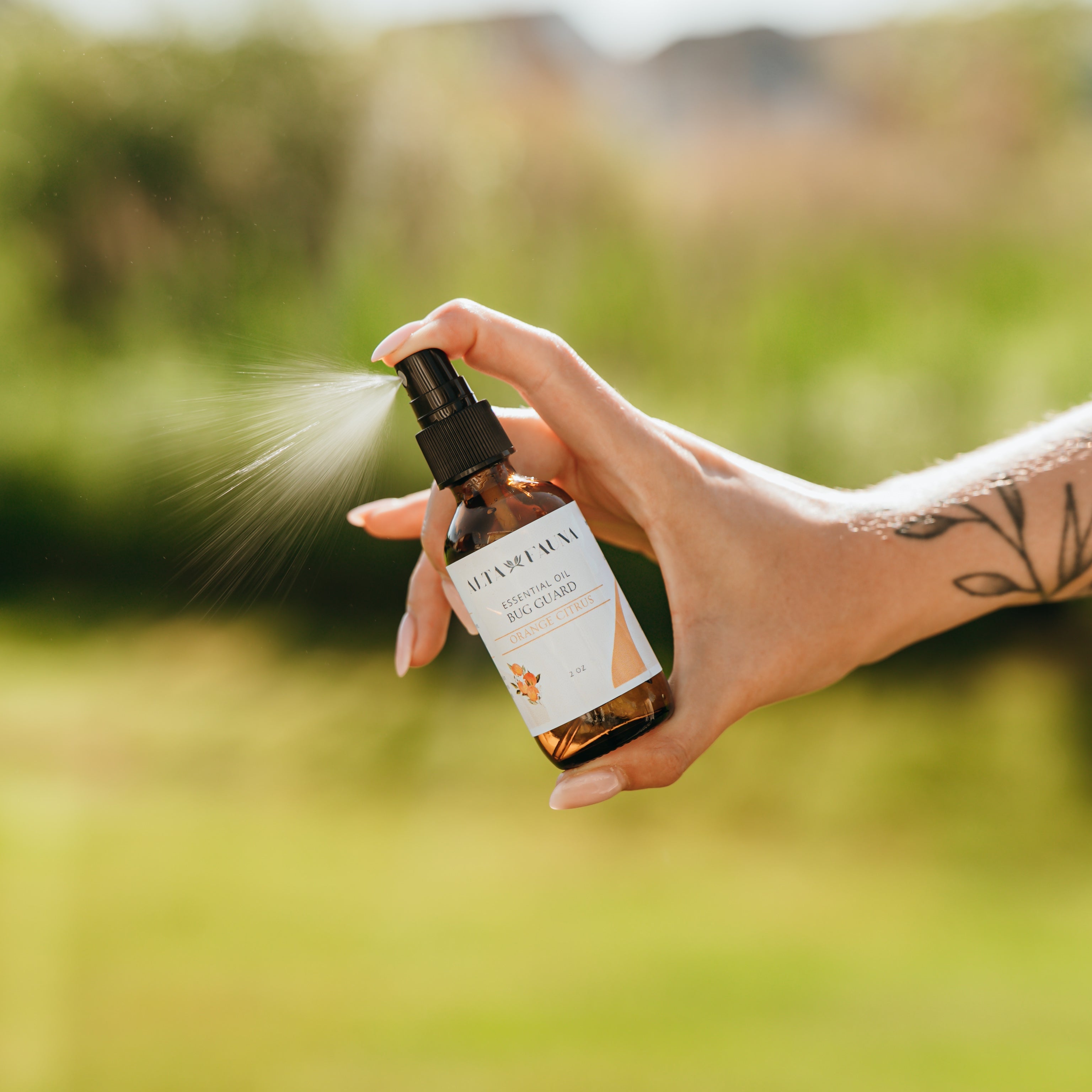 Alta Fauna Orange Citrus Bug Guard being held by a hand in an outdoor setting with a fine mist coming from the amber, glass spray bottle.