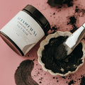 Charcoal and clay face mask jar next to a dish with moist product and a brush on a pink background.