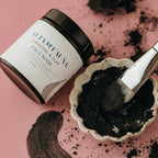 Charcoal and clay face mask jar next to a dish with moist product and a brush on a pink background.