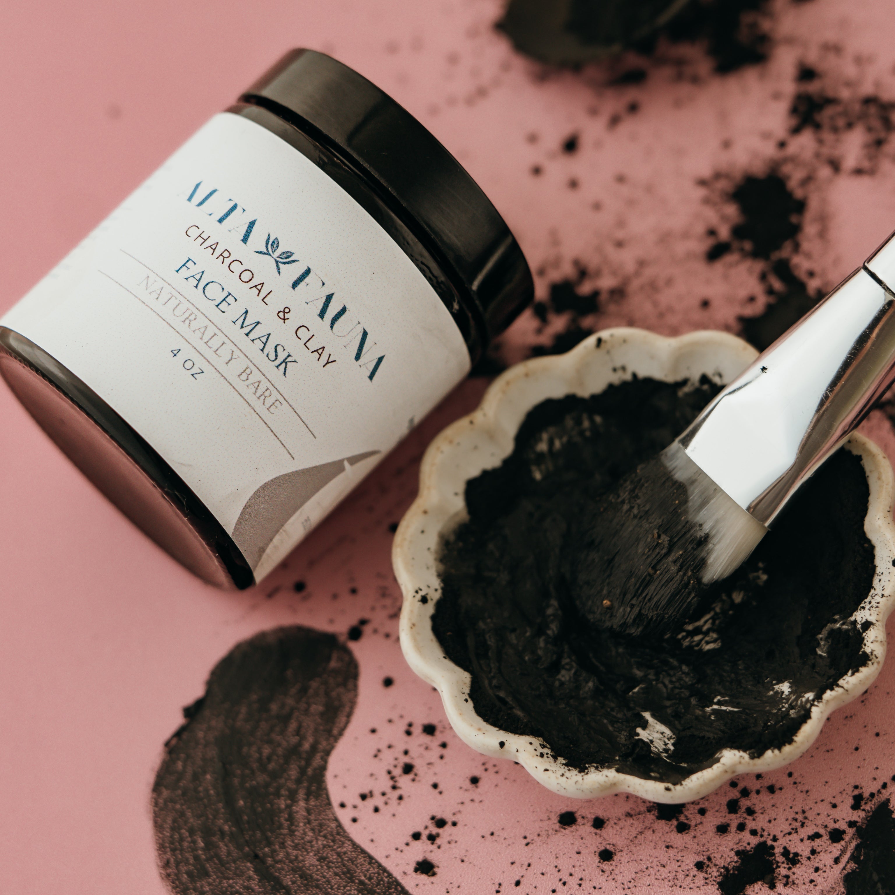 Charcoal and clay face mask jar next to a dish with moist product and a brush on a pink background.