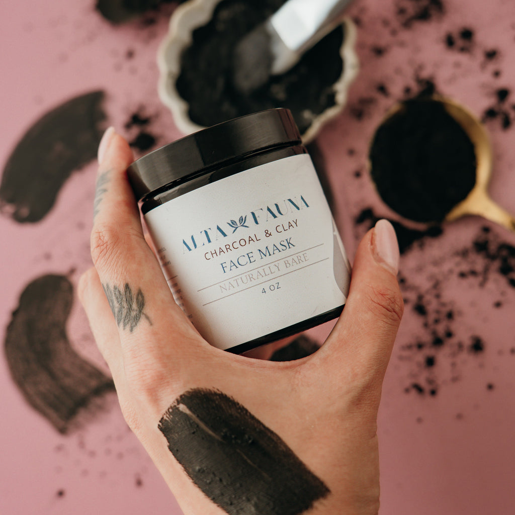 Hand holding a jar of Alta Fauna charcoal and clay face mask with black mask on hand against a pink background