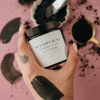 Hand holding a jar of Alta Fauna charcoal and clay face mask with black mask on hand against a pink background