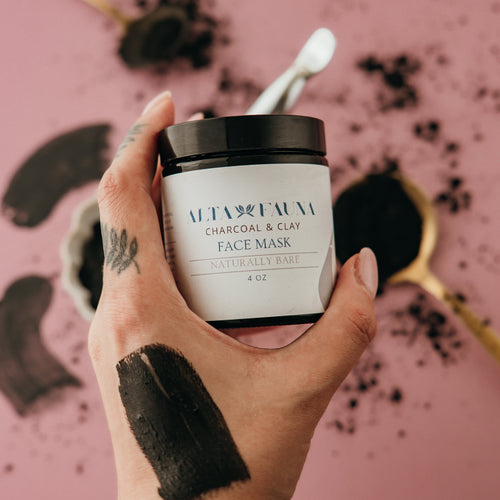 Hand holding a jar of Alta Fauna Charcoal & Clay Face Mask against a pink background with makeup tools.