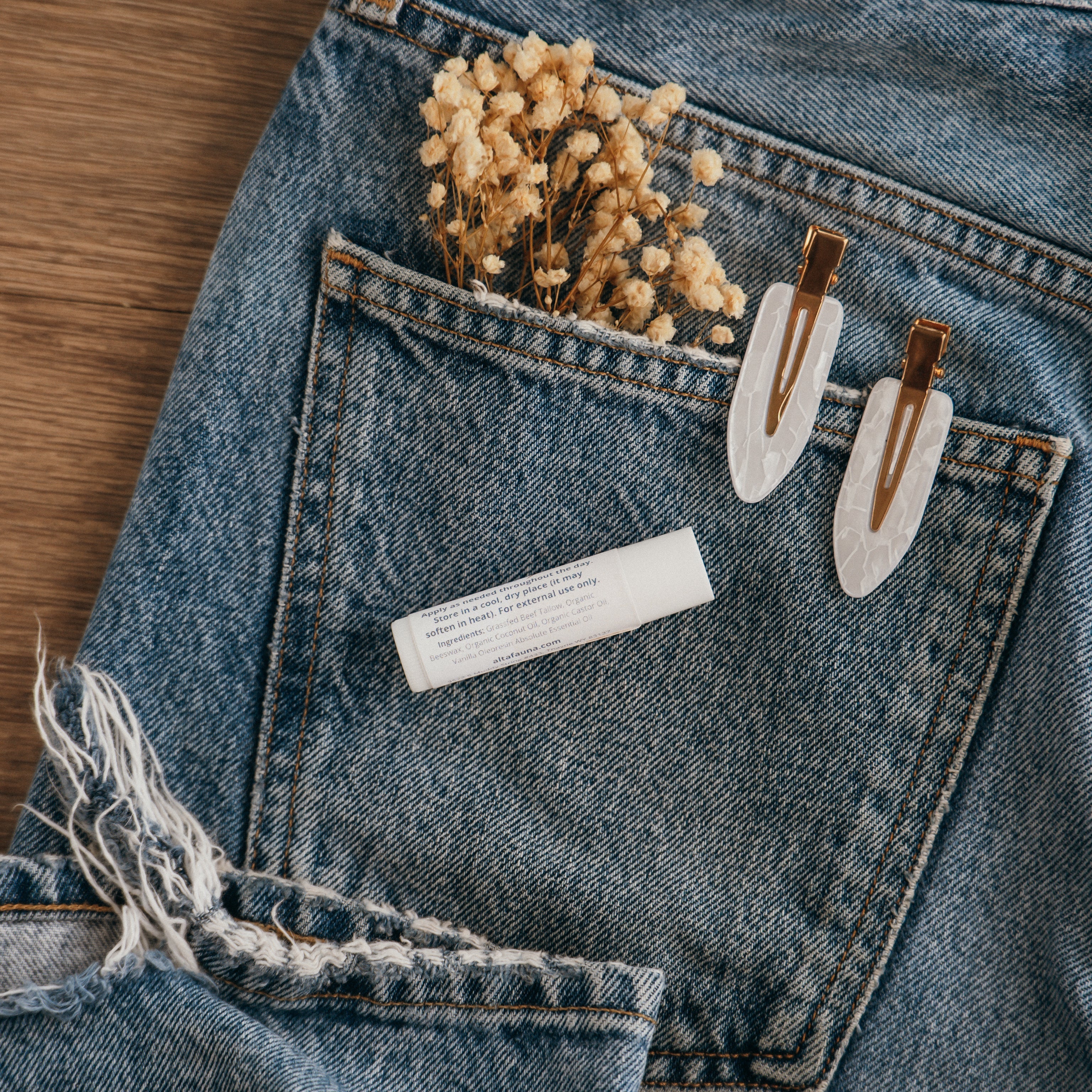 Close-up of Alta Fauna Lip balm packaging on a pocket on blue denim jeans with dried flowers, hair clips. Photo shows the back of the lip balm tube and includes ingredients. Alta Fauna lip balm styled with botanicals for small‑batch, chemical‑free skincare aesthetic