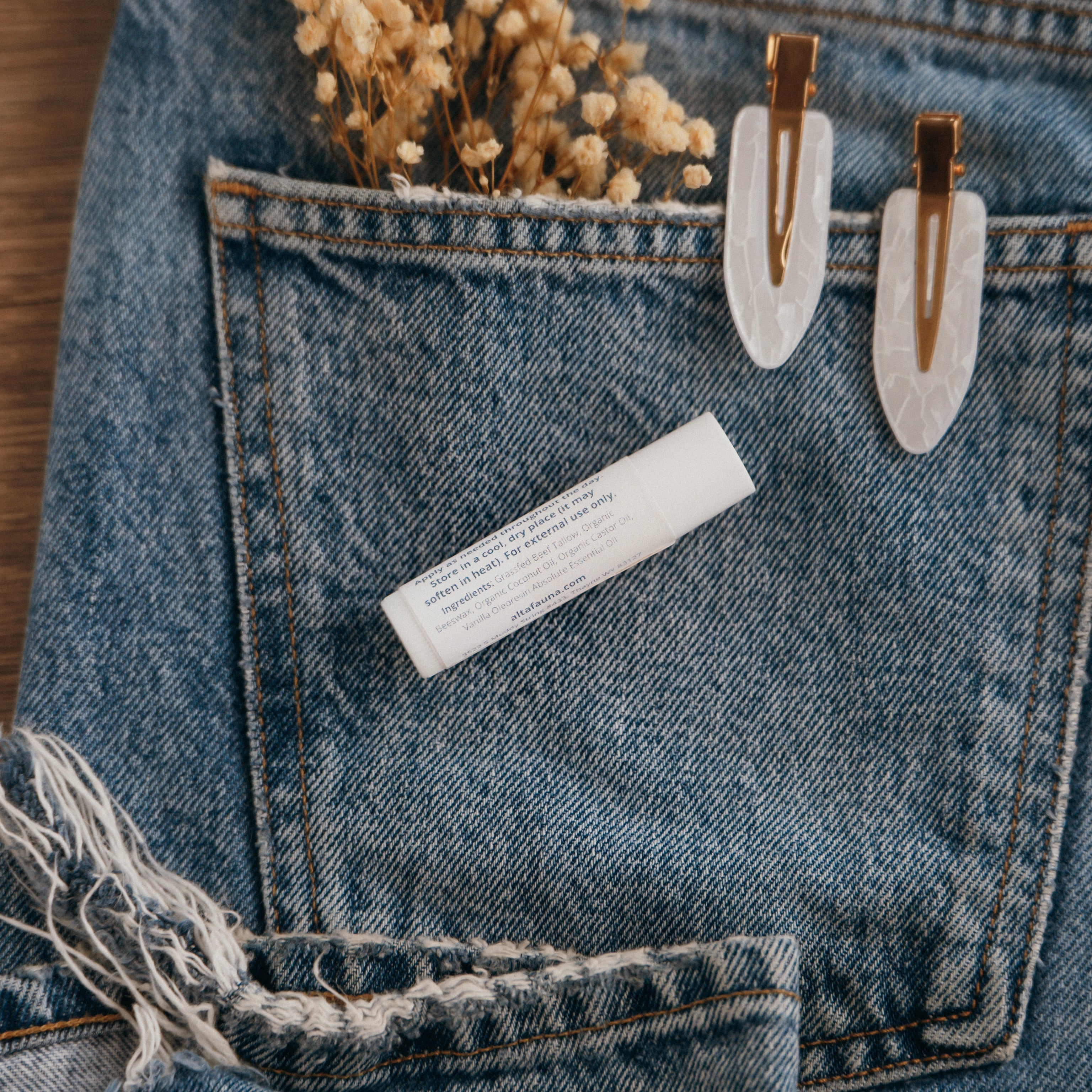Close-up of Alta Fauna Lip balm packaging on a pocket on blue denim jeans with dried flowers, hair clips. Photo shows the back of the lip balm tube and includes ingredients. Alta Fauna lip balm styled with botanicals for small‑batch, chemical‑free skincare aesthetic