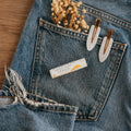 Close-up of Alta Fauna Banana Lip balm on a pocket on blue denim jeans with dried flowers, hair clips. Alta Fauna lip balm styled with botanicals for small‑batch, chemical‑free skincare.