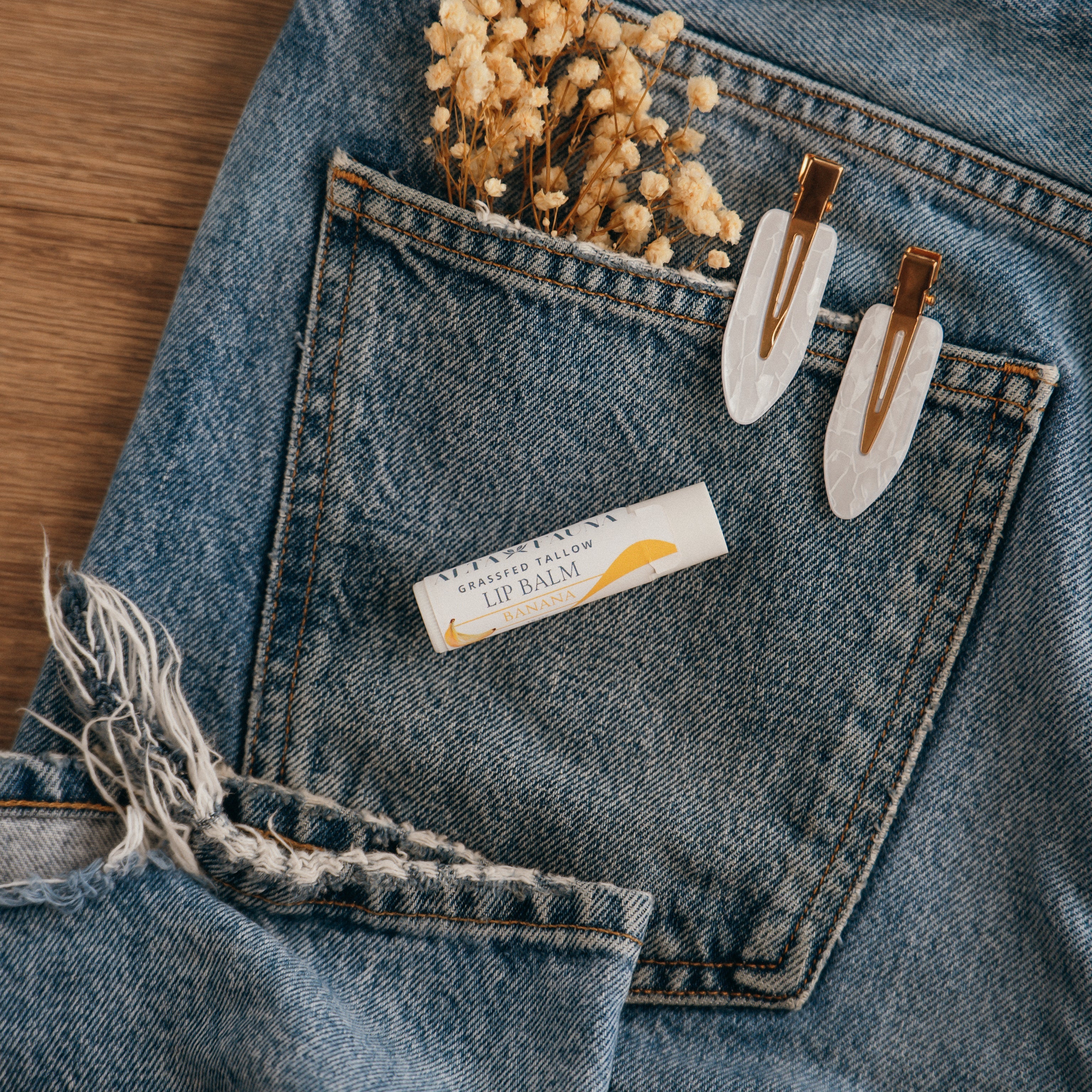 Close-up of Alta Fauna Banana Lip balm on a pocket on blue denim jeans with dried flowers, hair clips. Alta Fauna lip balm styled with botanicals for small‑batch, chemical‑free skincare.