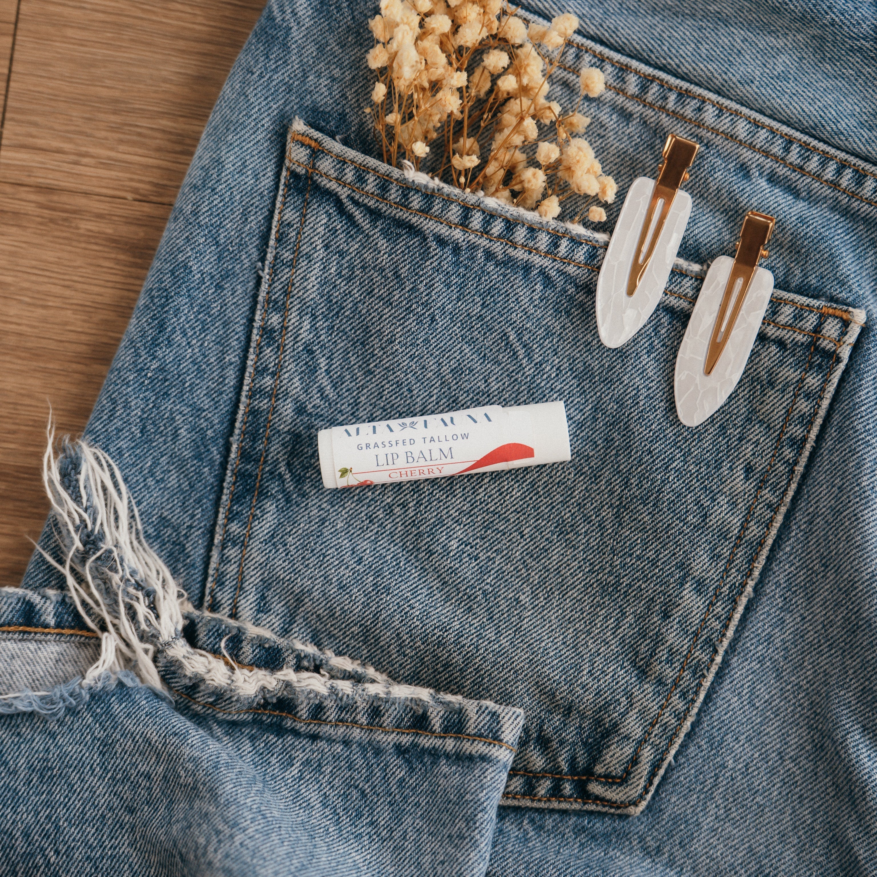 Close-up of Alta Fauna Cherry Lip balm on a pocket on blue denim jeans with dried flowers, hair clips. Alta Fauna lip balm styled with botanicals for small‑batch, chemical‑free skincare.