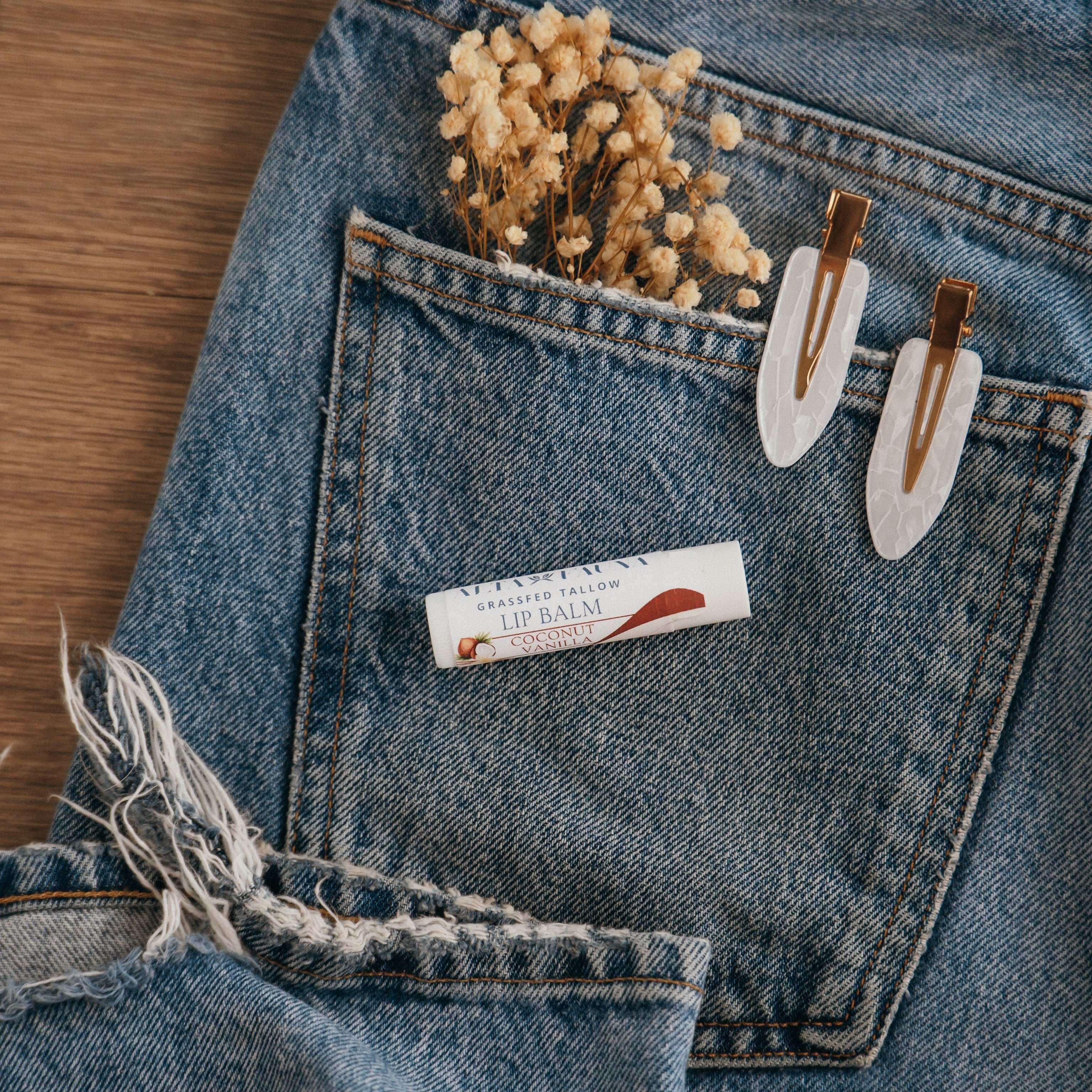 Close-up of Alta Fauna Coconut Vanilla Lip balm on a pocket on blue denim jeans with dried flowers, hair clips. Alta Fauna lip balm styled with botanicals for small‑batch, chemical‑free skincare.