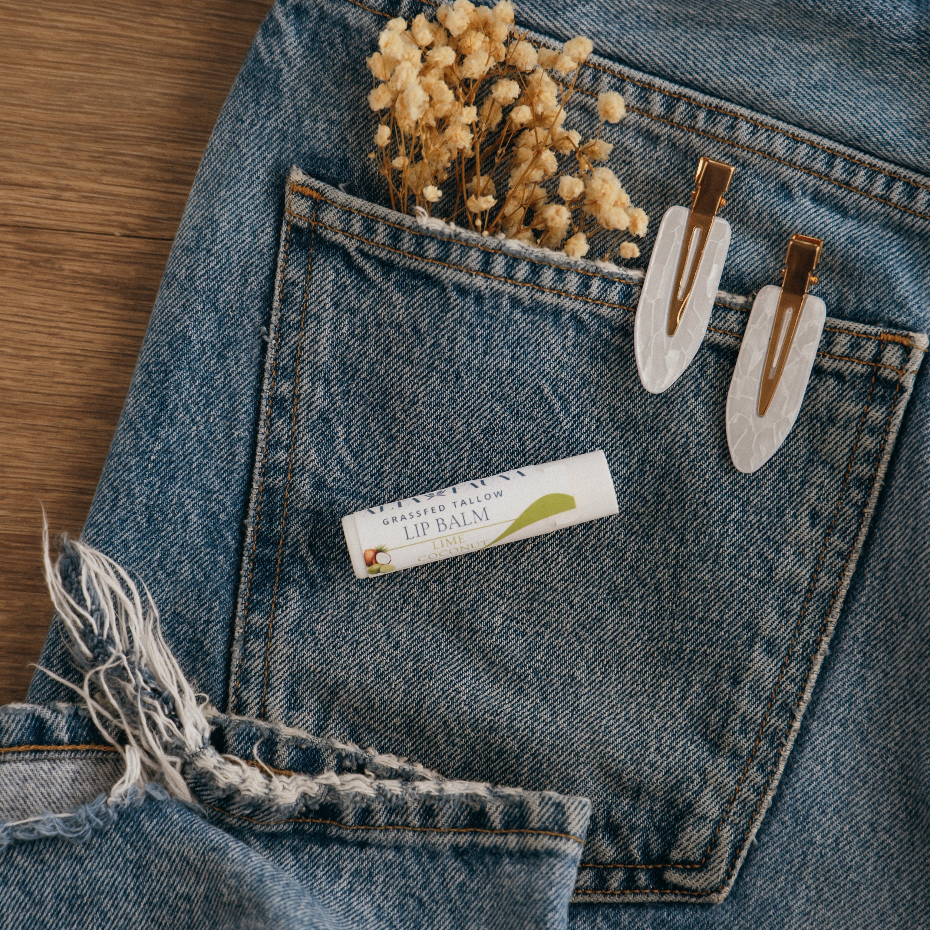 Close-up of Alta Fauna Lime Coconut Lip balm on a pocket on blue denim jeans with dried flowers, hair clips. Alta Fauna lip balm styled with botanicals for small‑batch, chemical‑free skincare.