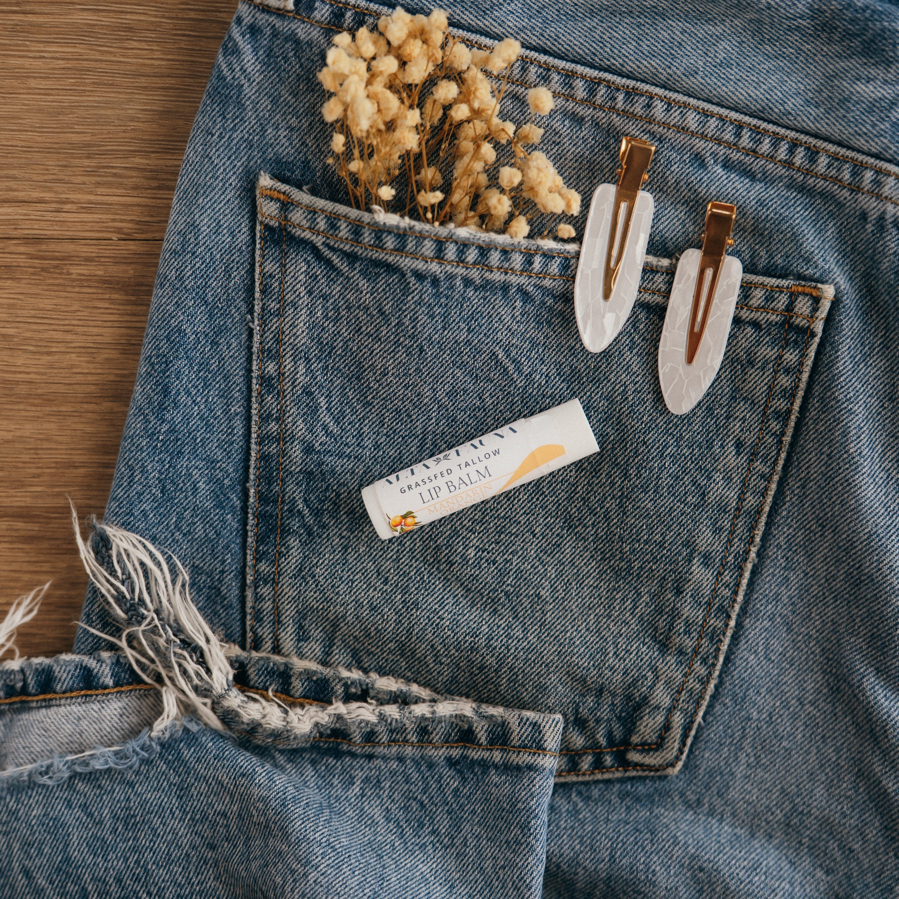 Close-up of Alta Fauna Mandarin Orange Lip balm on a pocket on blue denim jeans with dried flowers, hair clips. Alta Fauna lip balm styled with botanicals for small‑batch, chemical‑free skincare aesthetic