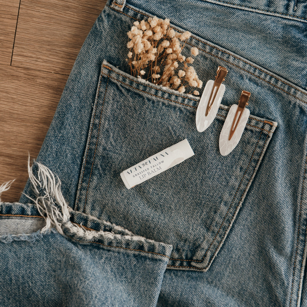 Close-up of Alta Fauna Naturally Bare Lip balm on a pocket on blue denim jeans with dried flowers, hair clips. Alta Fauna lip balm styled with botanicals for small‑batch, chemical‑free skincare.