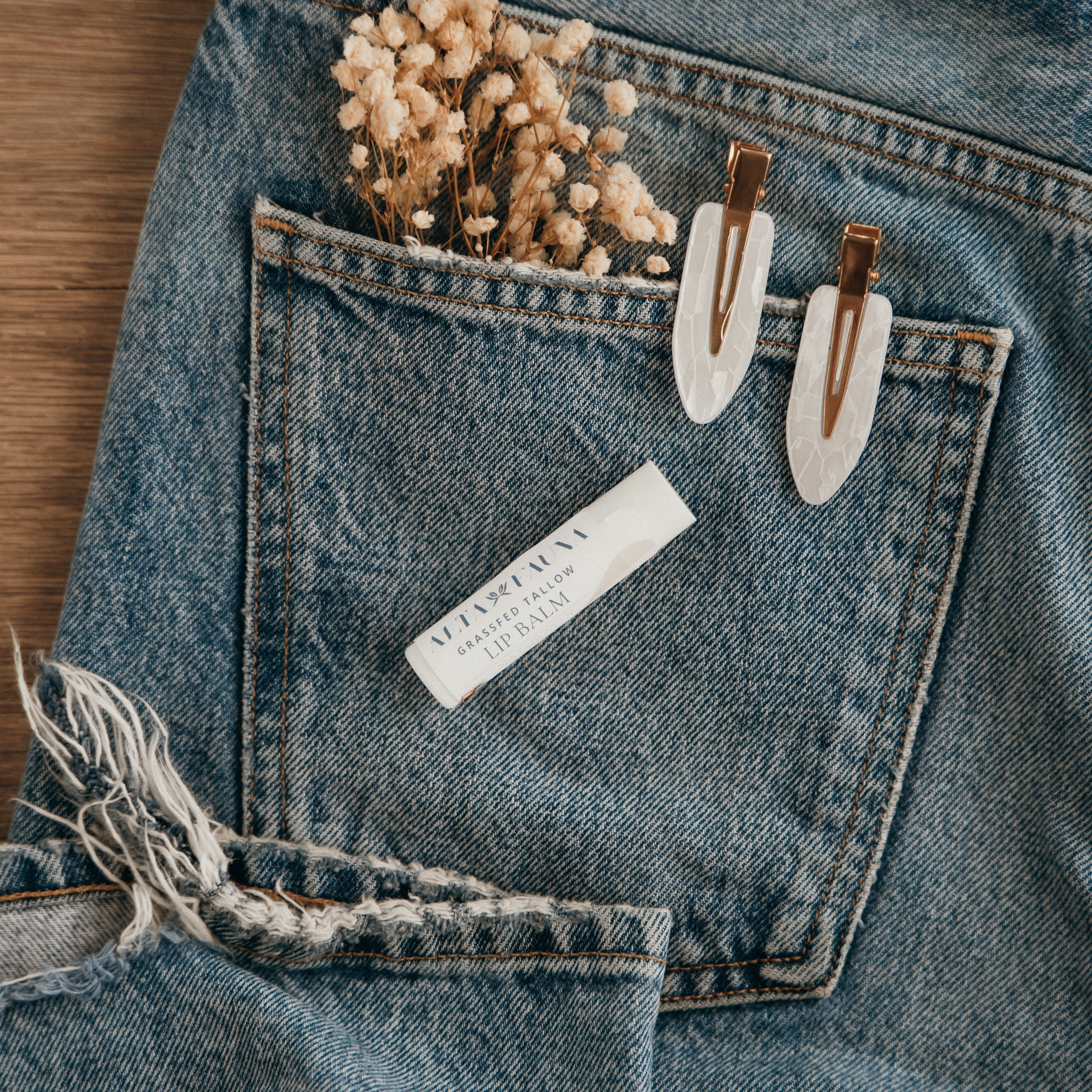 Close-up of Alta Fauna Naturally Bare Lip balm on a pocket on blue denim jeans with dried flowers, hair clips. Alta Fauna lip balm styled with botanicals for small‑batch, chemical‑free skincare.
