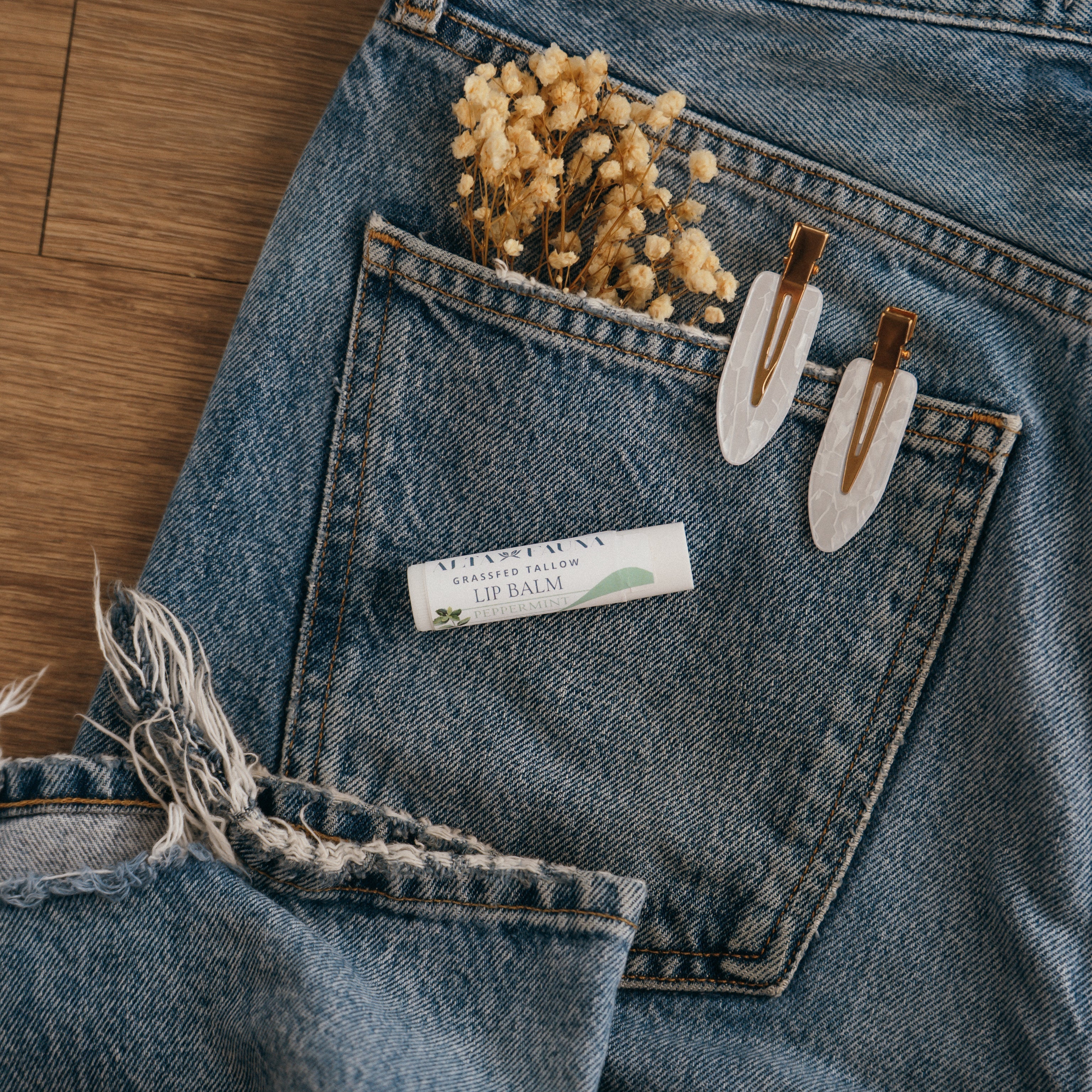 Close-up of Alta Fauna Peppermint Lip balm on a pocket on blue denim jeans with dried flowers, hair clips. Alta Fauna lip balm styled with botanicals for small‑batch, chemical‑free skincare.