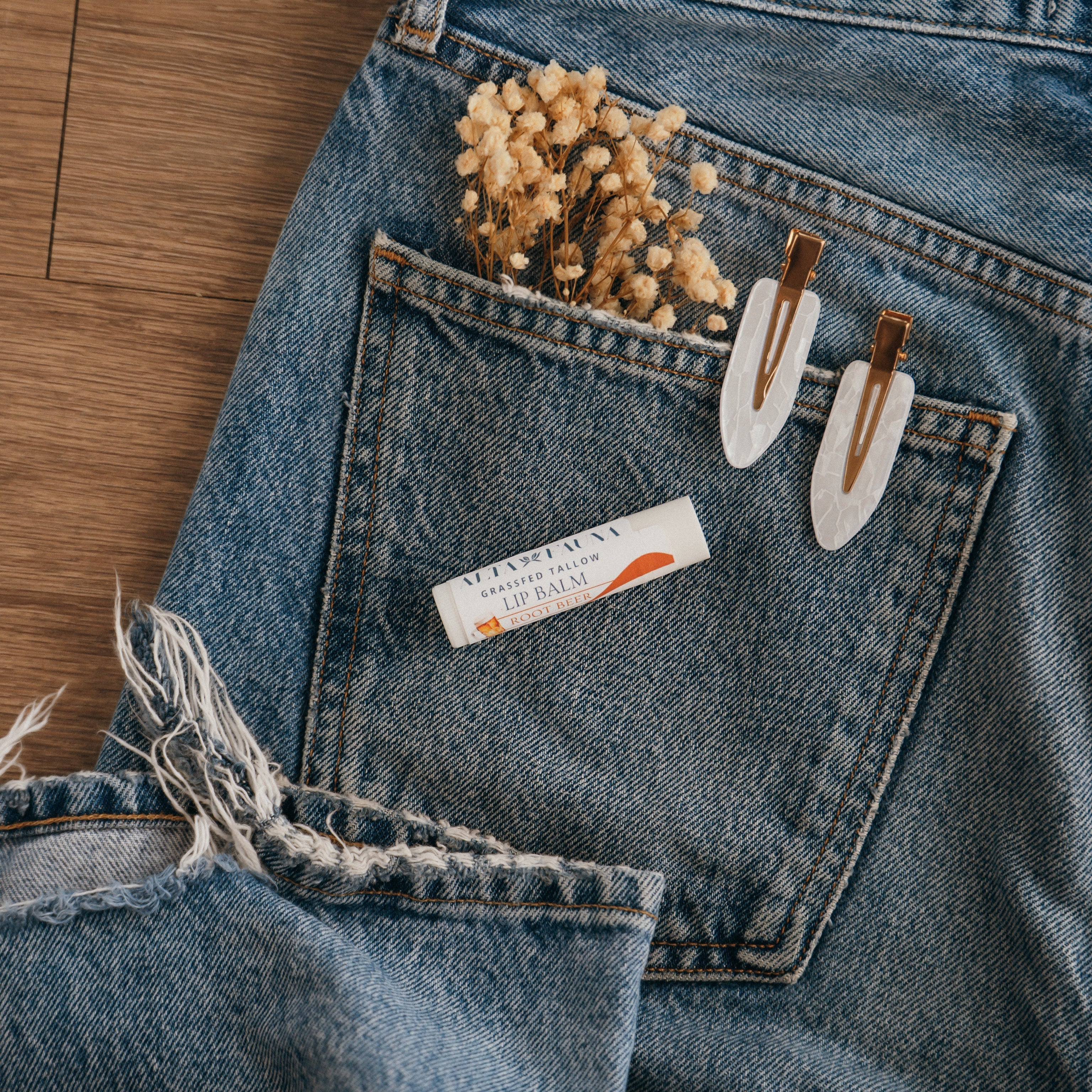 Close-up of Alta Fauna Root beer Lip balm on a pocket on blue denim jeans with dried flowers, hair clips. Alta Fauna lip balm styled with botanicals for small‑batch, chemical‑free skincare.