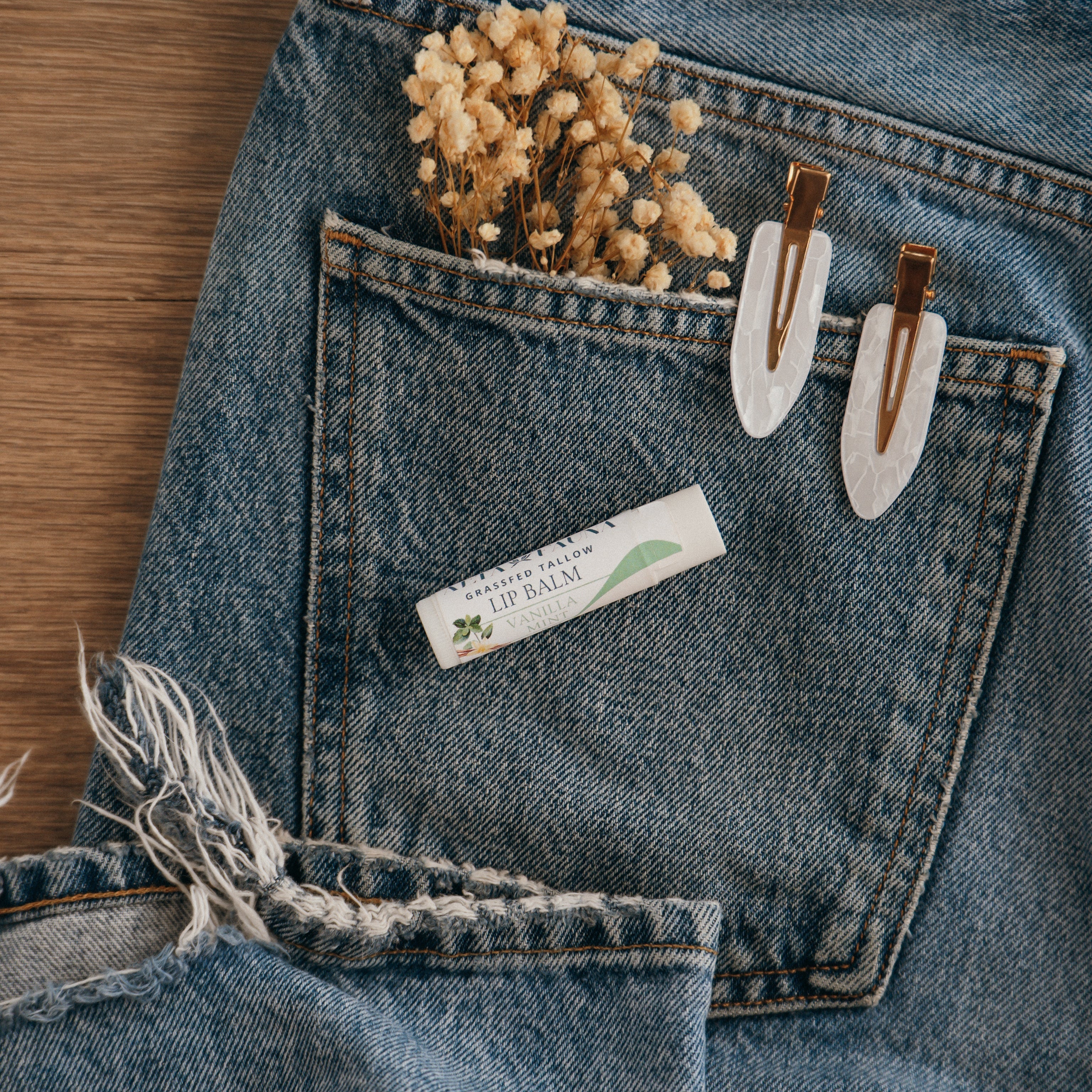 Close-up of Alta Fauna Vanilla Mint Lip balm on a pocket on blue denim jeans with dried flowers, hair clips. Alta Fauna lip balm styled with botanicals for small‑batch, chemical‑free skincare.