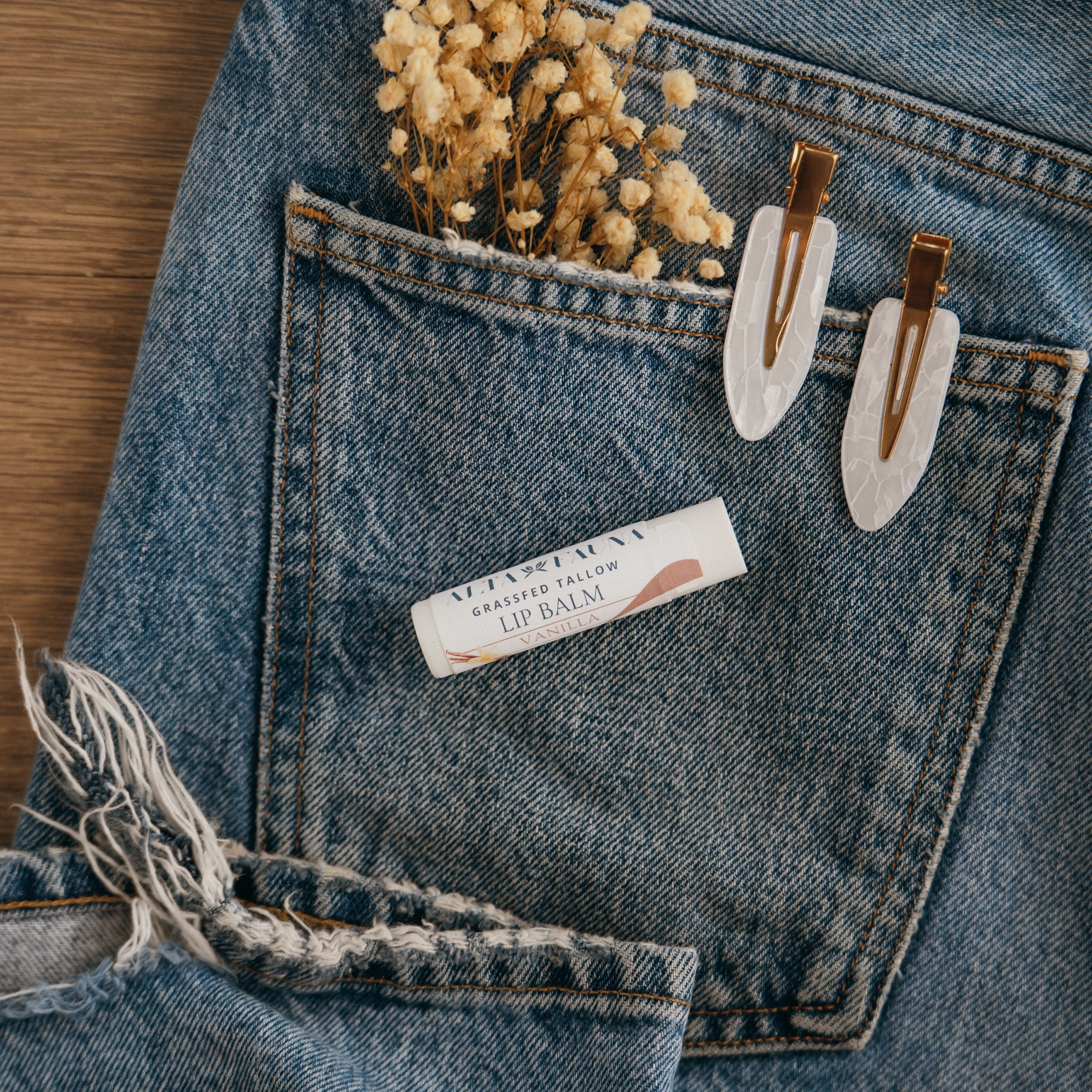 Close-up of Alta Fauna Vanilla Lip balm on a pocket on blue denim jeans with dried flowers, hair clips. Alta Fauna lip balm styled with botanicals for small‑batch, chemical‑free skincare.