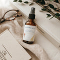 Bottle of Alta Fauna Room & Linen spray in Cedarwood on a textured surface with books and glasses in the background. Natural room and linen spray for home fragrance with clean, simple ingredients.