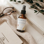 Bottle of Alta Fauna Room & Linen spray in Cedarwood on a textured surface with books and glasses in the background. Natural room and linen spray for home fragrance with clean, simple ingredients.