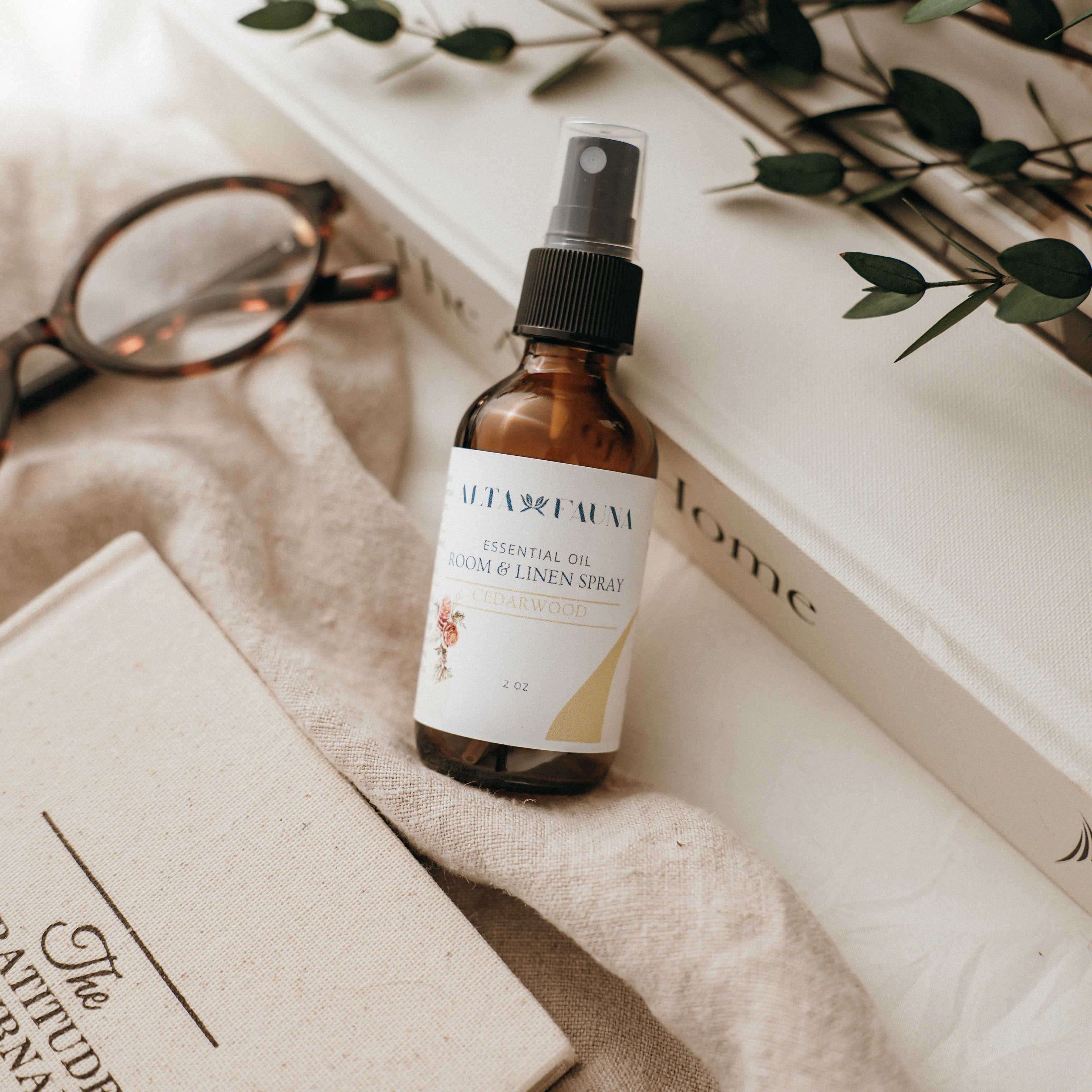 Bottle of Alta Fauna Room & Linen spray in Cedarwood on a textured surface with books and glasses in the background. Natural room and linen spray for home fragrance with clean, simple ingredients.