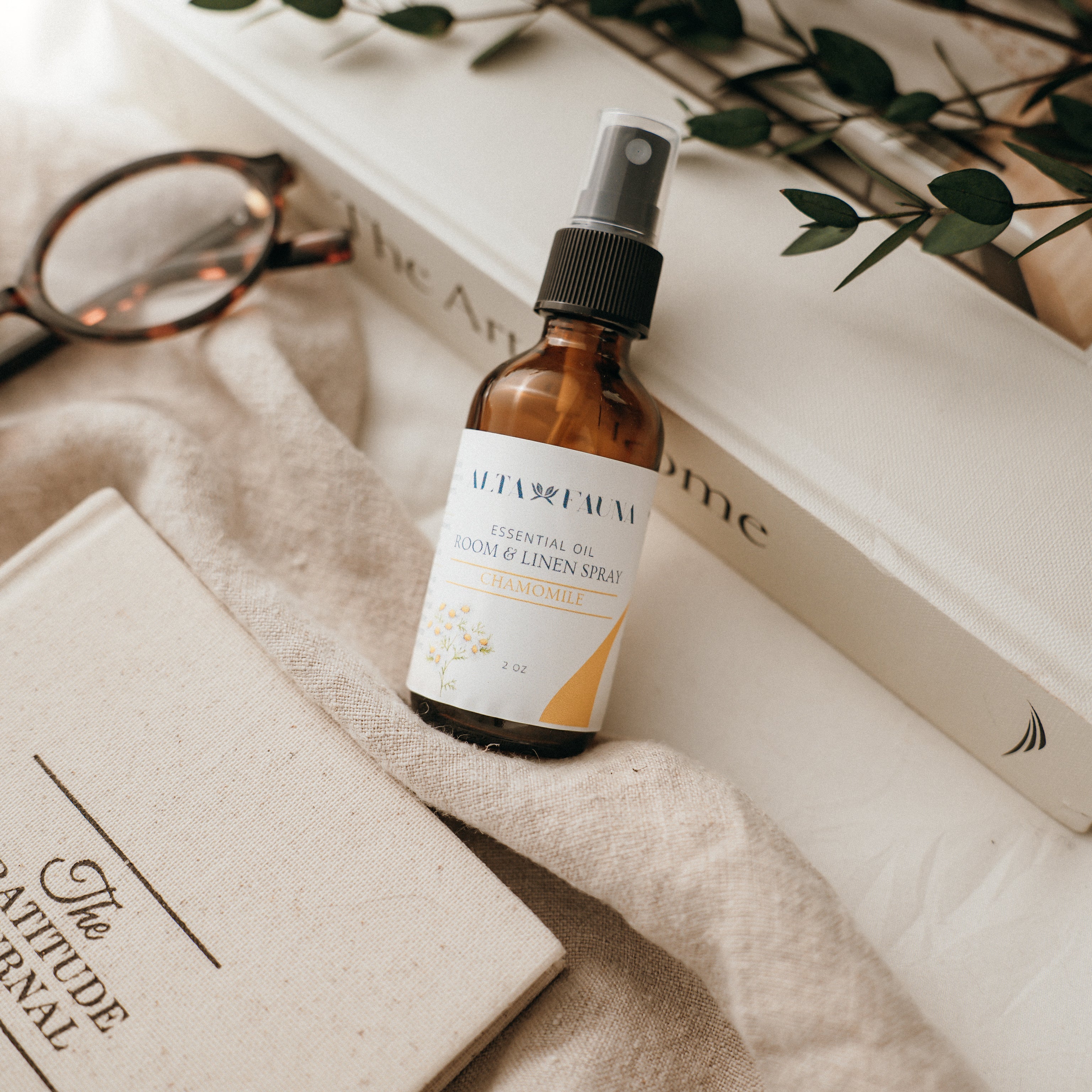 Bottle of Alta Fauna Room & Linen spray in Chamomile on a textured surface with books and glasses in the background. Natural room and linen spray for home fragrance with clean, simple ingredients.