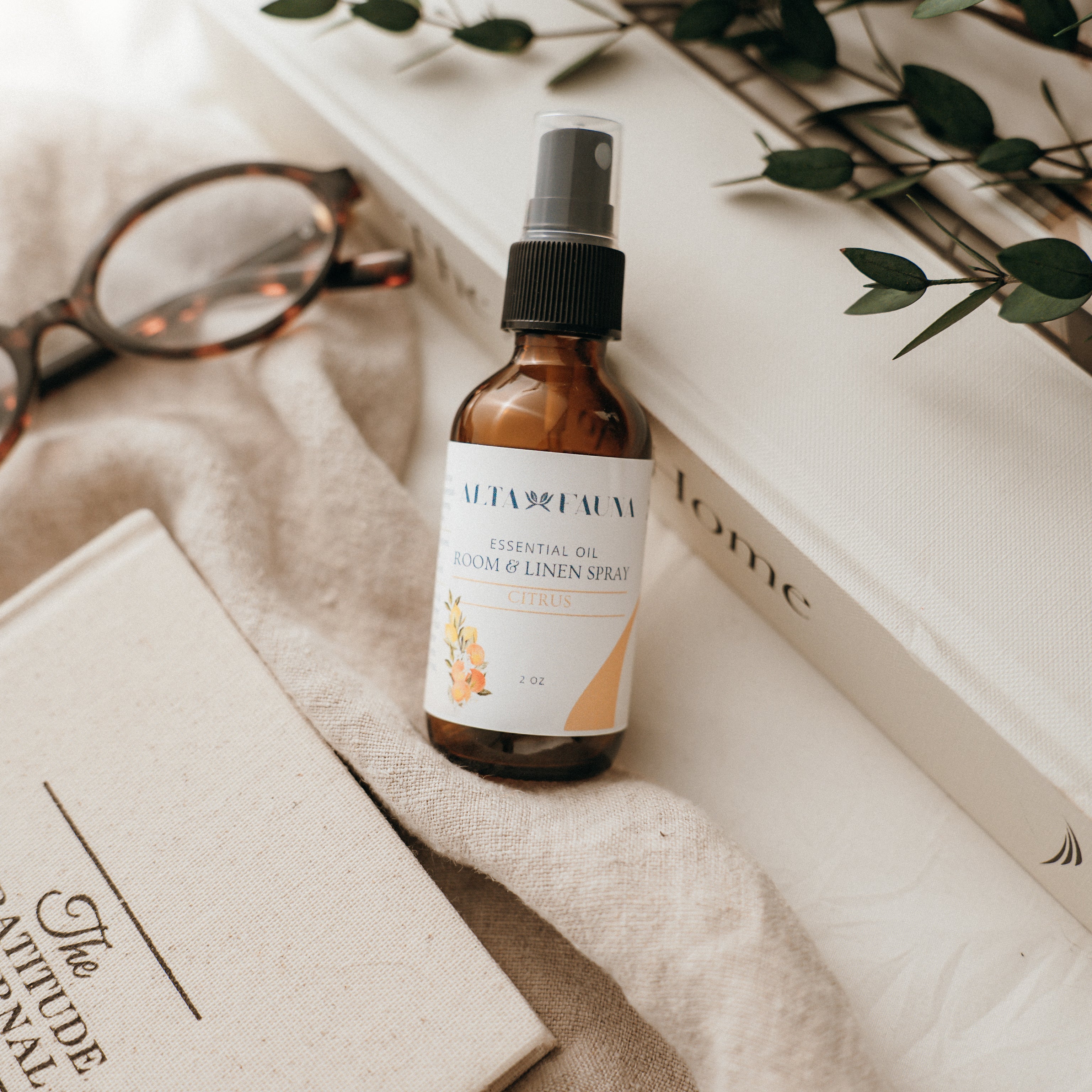 Bottle of Alta Fauna Room & Linen spray in Citrus on a textured surface with books and glasses in the background. Natural room and linen spray for home fragrance with clean, simple ingredients.