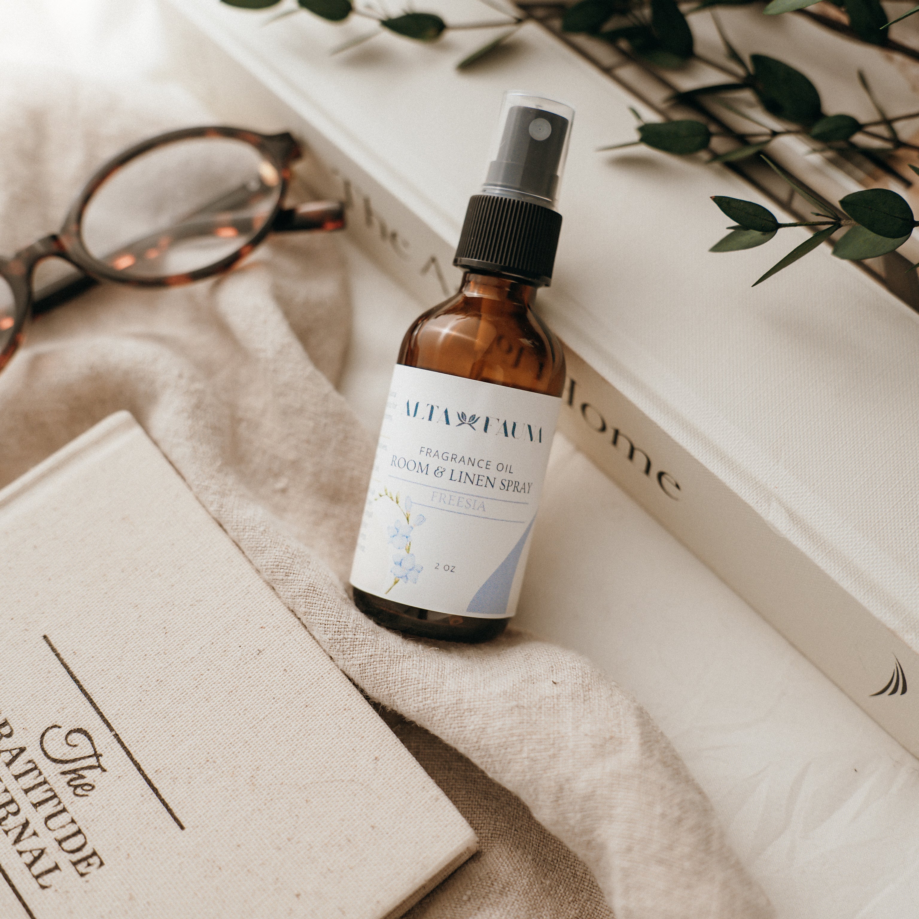 Bottle of Alta Fauna Room & Linen spray in Freesia on a textured surface with books and glasses in the background. Natural room and linen spray for home fragrance with clean, simple ingredients.