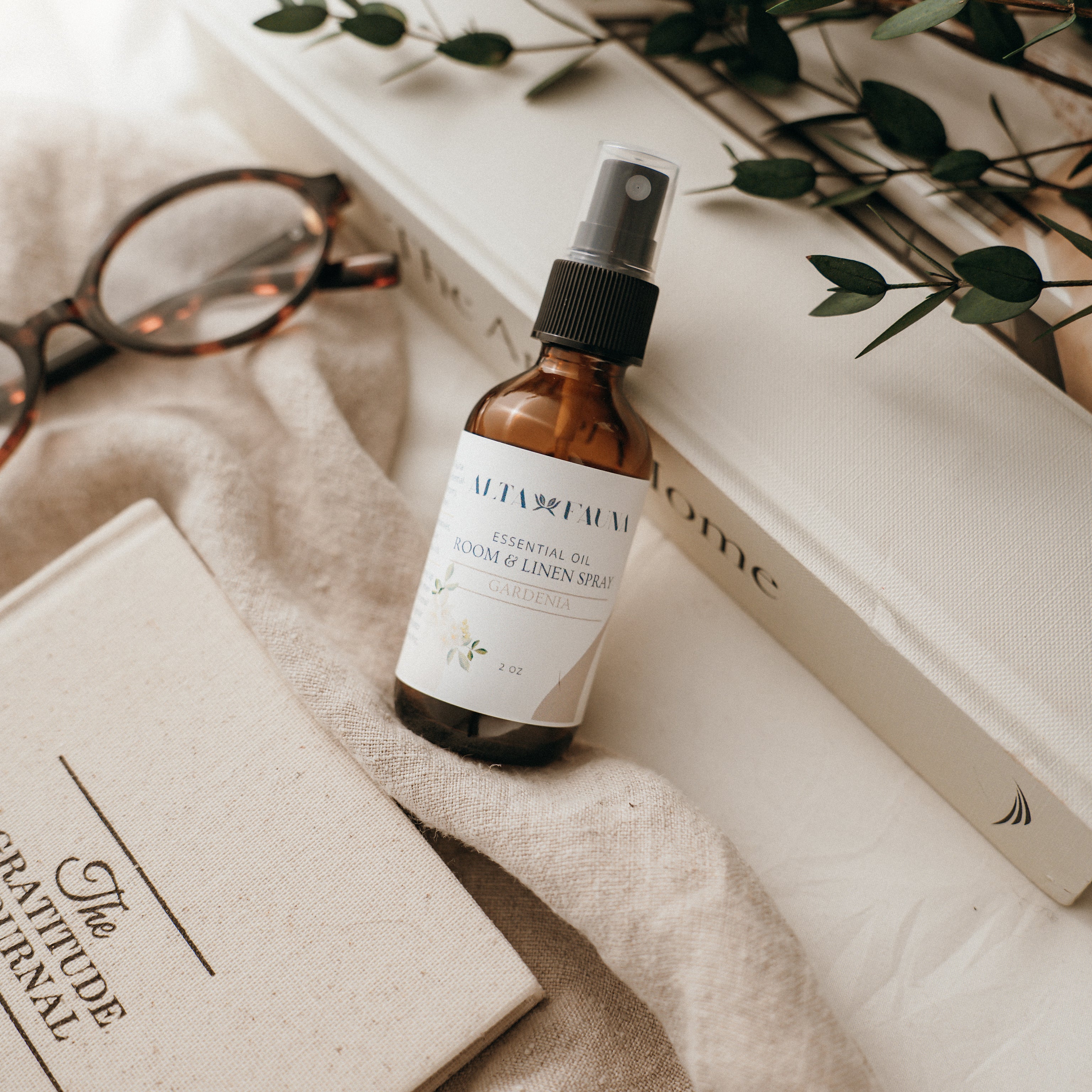 Bottle of Alta Fauna Room & Linen spray in Gardenia on a textured surface with books and glasses in the background. Natural room and linen spray for home fragrance with clean, simple ingredients.