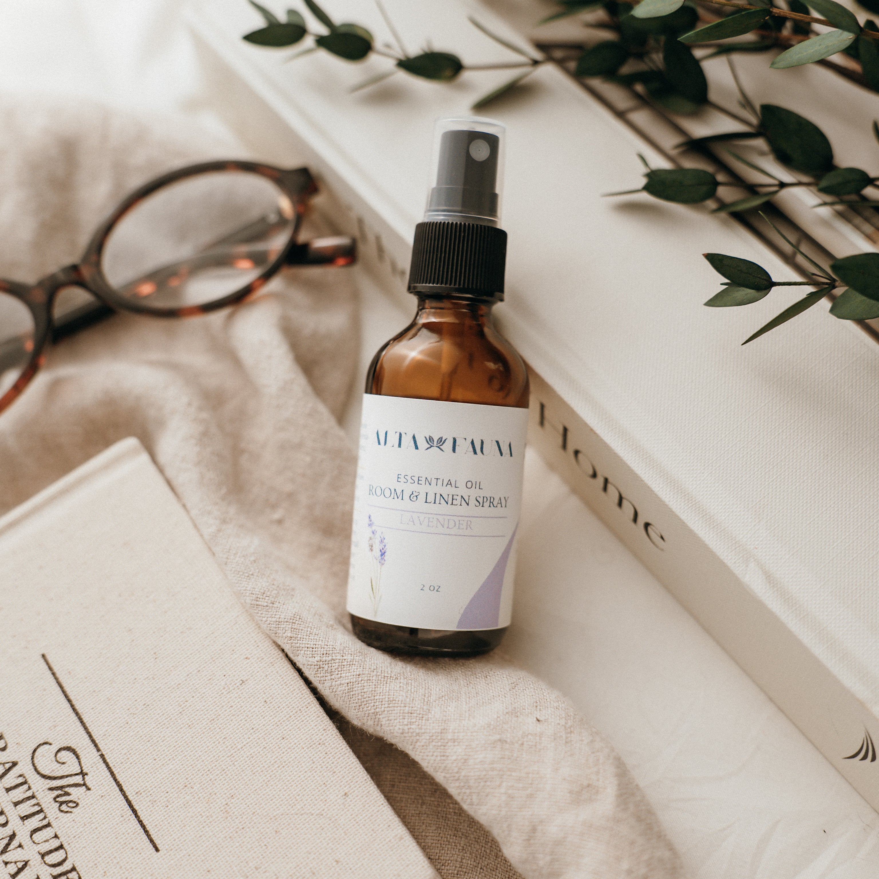 Bottle of Alta Fauna Room & Linen spray in Lavender on a textured surface with books and glasses in the background. Natural room and linen spray for home fragrance with clean, simple ingredients.