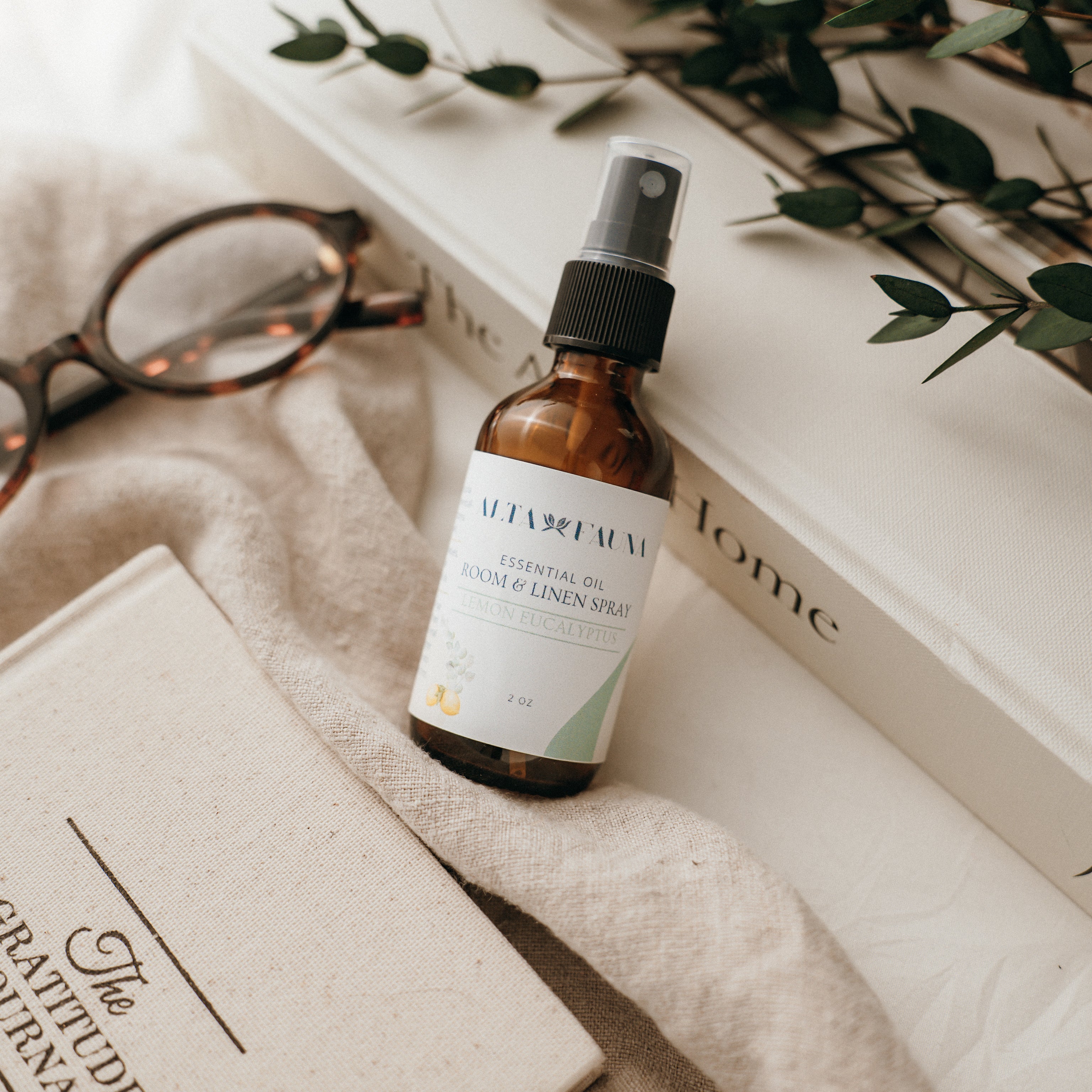 Bottle of Alta Fauna Room & Linen spray in Lemon Eucalyptus on a textured surface with books and glasses in the background. Natural room and linen spray for home fragrance with clean, simple ingredients.