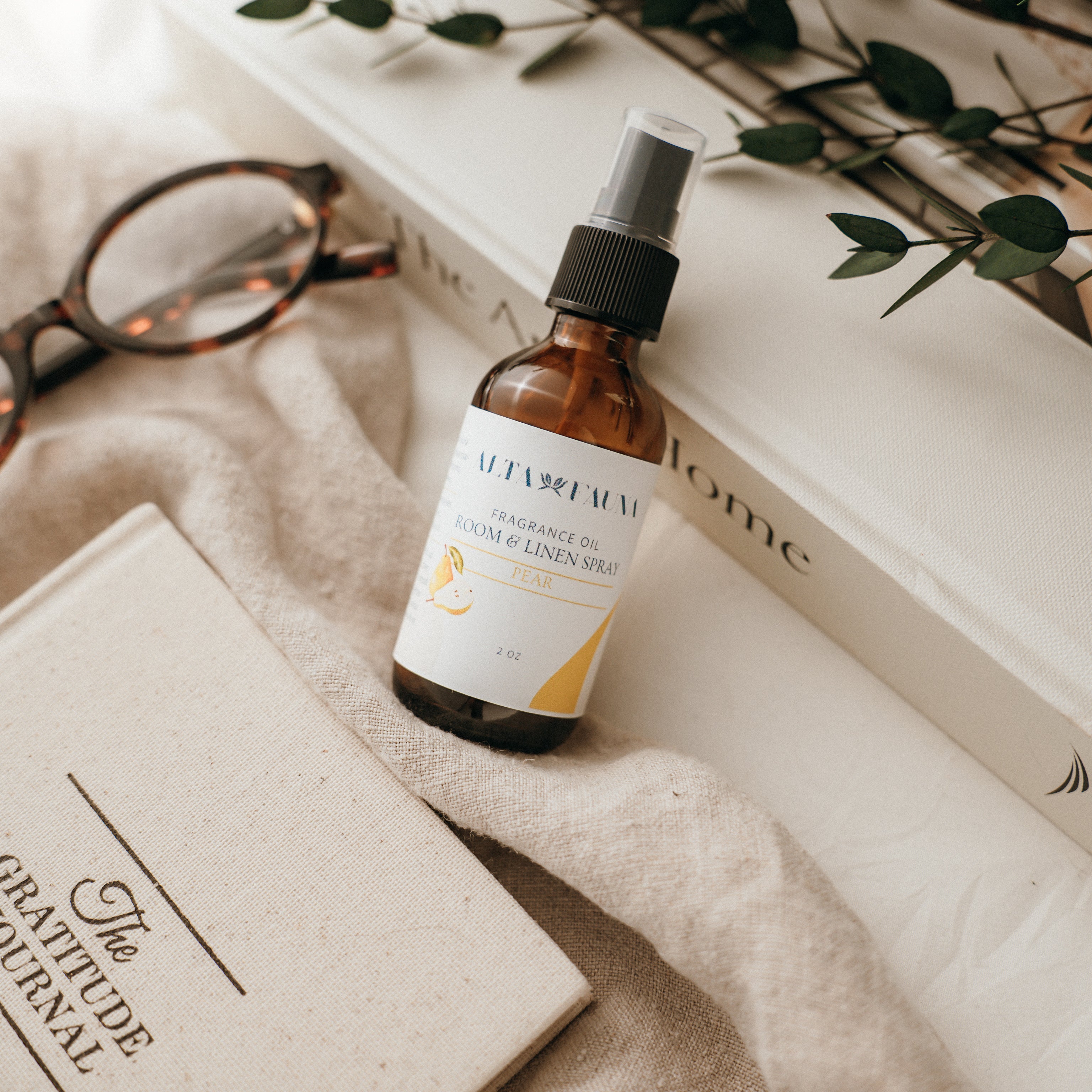 Bottle of Alta Fauna Room & Linen spray in Pear on a textured surface with books and glasses in the background. Natural room and linen spray for home fragrance with clean, simple ingredients.
