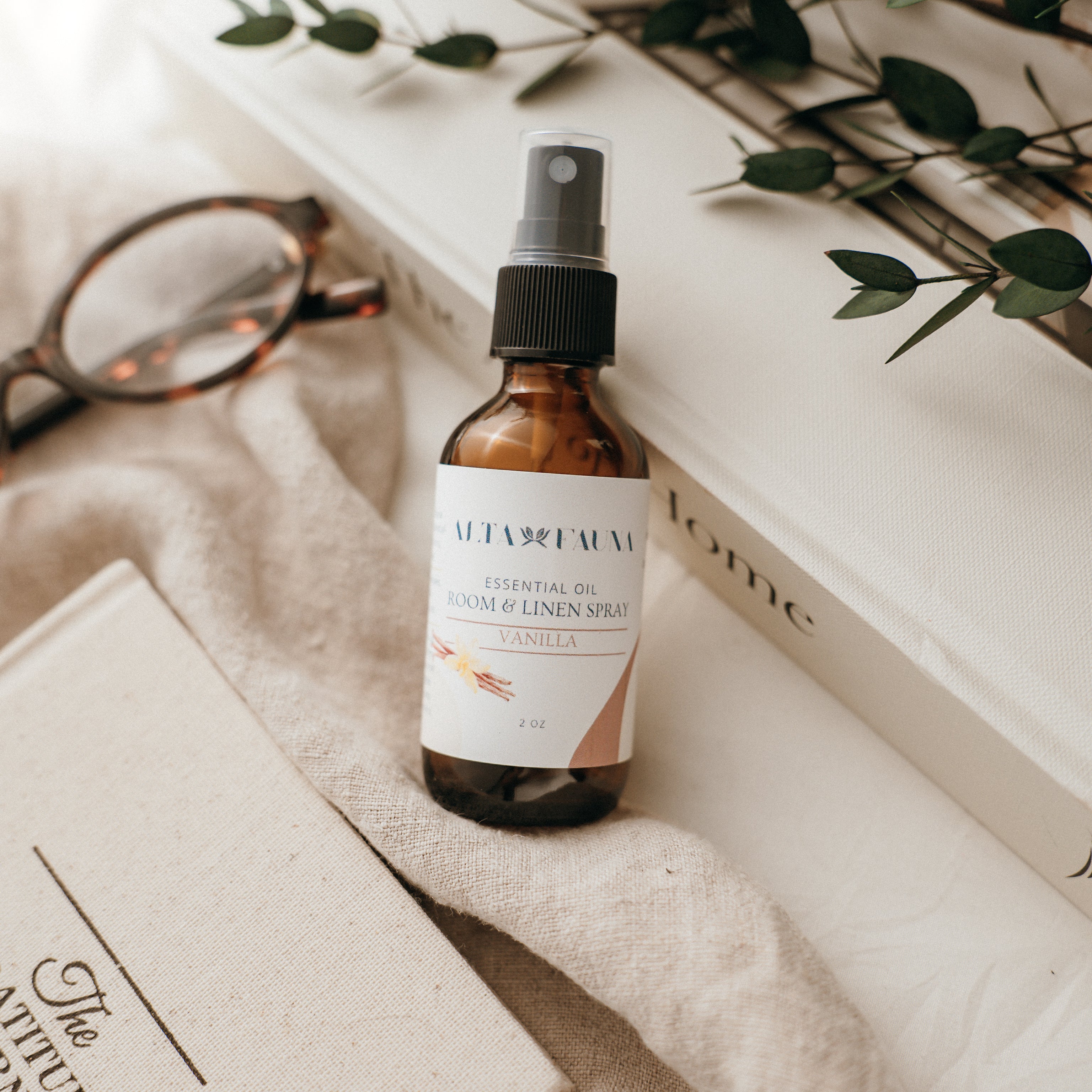 Bottle of Alta Fauna Room & Linen spray in Vanilla on a textured surface with books and glasses in the background. Natural room and linen spray for home fragrance with clean, simple ingredients.