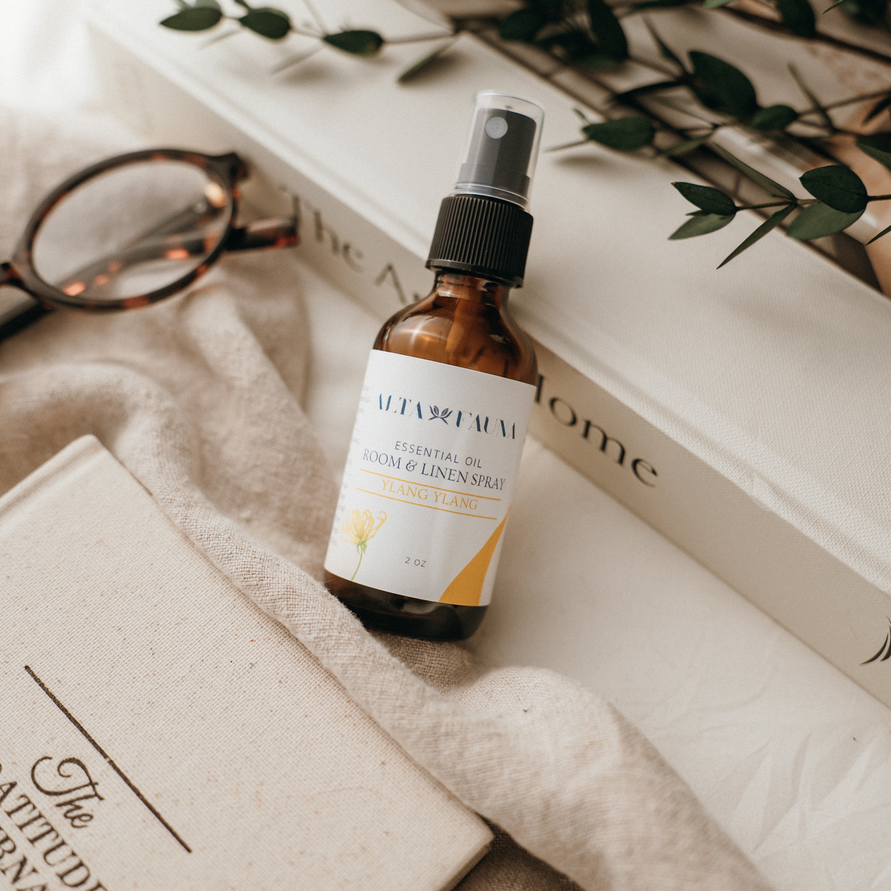 Bottle of Alta Fauna Room & Linen spray in Ylang Ylang on a textured surface with books and glasses in the background. Natural room and linen spray for home fragrance with clean, simple ingredients.