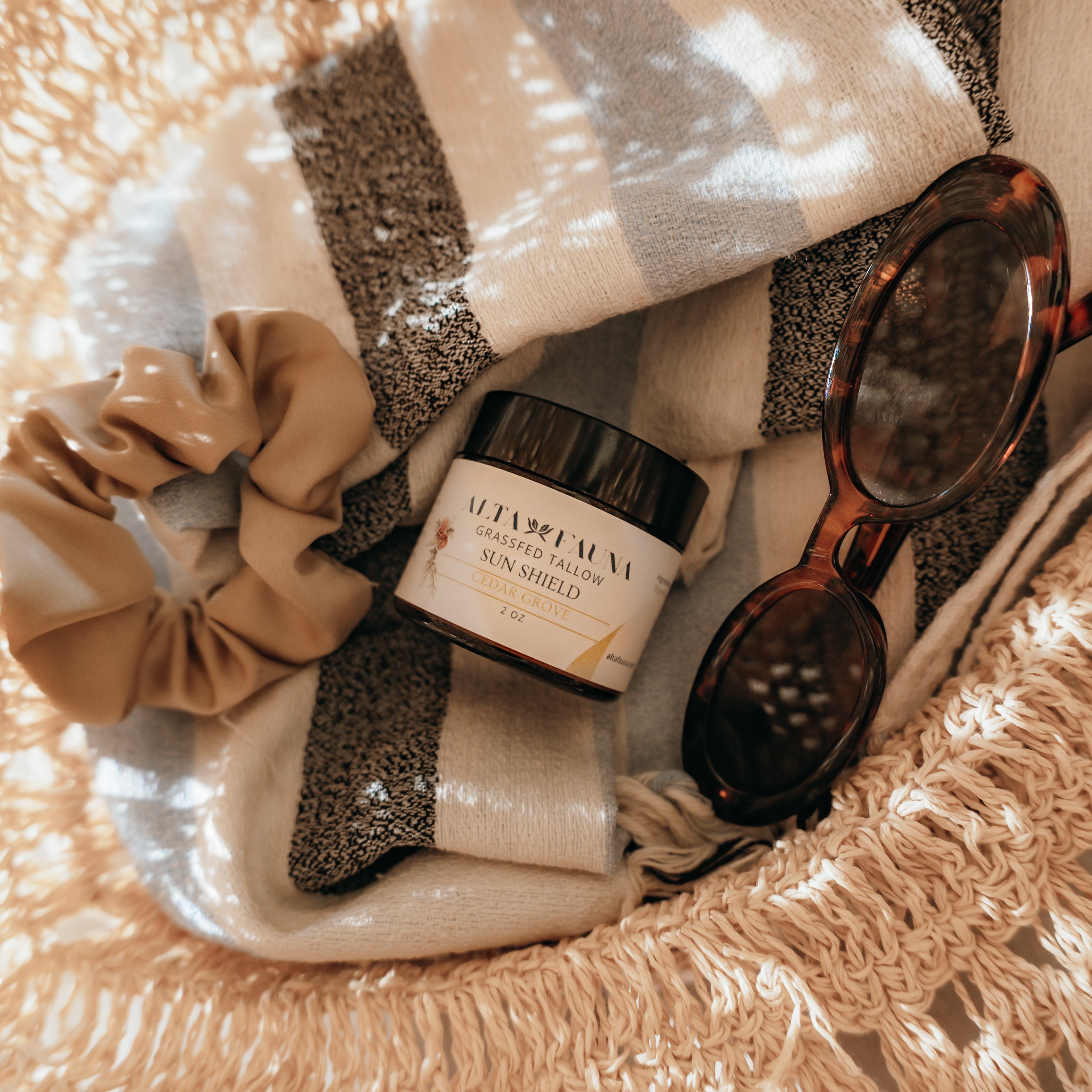 Basket with Alta Fauna Sun Shield lotion, scrunchie, and sunglasses on a beach towel.