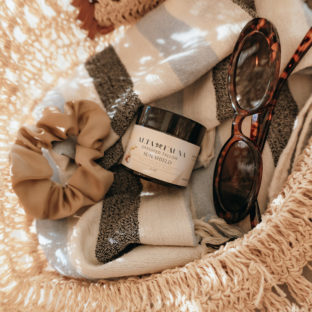 Cosmetic bag with Alta Fauna Sun Shield lotion, scrunchie, and sunglasses on a beach towel.